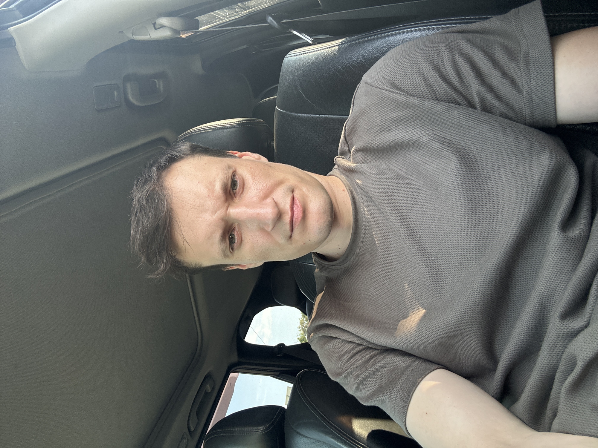 A mid-shot, selfie-style photograph shows a man sitting in the driver's seat of a car, looking directly at the camera. The man has short, dark hair and is wearing a grey t-shirt. Sunlight streams into the car from the right side of the frame, illuminating his face and upper body. The interior of the car, including the dashboard and seats, is visible. The man's expression is neutral, with a slight frown. The background shows the open car door and a glimpse of the sky and trees outside, suggesting an outdoor location. The specific location in Shatava, Ukraine, cannot be determined from the visual information provided.