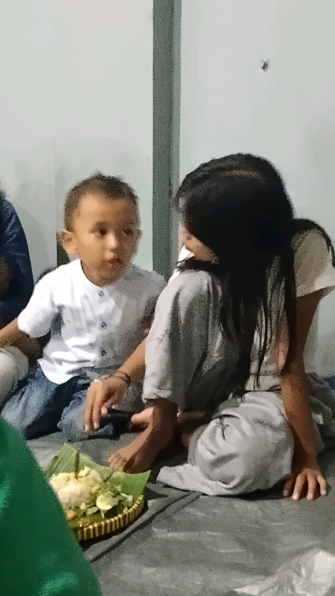 Two children are sitting on a mat on the floor. The child on the left is a boy with short dark hair, wearing a white button-up shirt and blue jeans. He is looking to the right with his mouth slightly open. The child on the right is a girl with long black hair, wearing a grey t-shirt and grey pants. She is facing away from the camera, looking towards the boy. Their hands are touching. In front of them, on a woven mat lined with banana leaves, is a serving of food that appears to include rice, some vegetables, and what looks like cucumber slices. The background is a plain wall, with a vertical grey beam on the right. The lighting is somewhat dim and diffused. The image appears to have been taken indoors, possibly in a home in Randuagung, Indonesia.