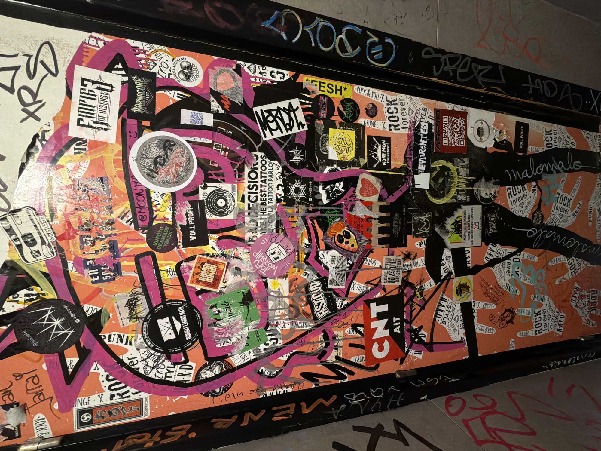 The image depicts a sticker-covered surface, possibly a door or panel, in an urban environment. The stickers vary in size, color, and design, featuring band names ("Empires of Disease," "BarnaCORE fest"), phrases ("Rock & Roll," "Grunge X"), and logos. Black and pink graffiti-style markings are layered over the stickers. The location is likely Barcelona, Spain, based on the sticker text, which mentions "Jardines de Hacia, Barcelona" and the Catalan word "BUENDATO." The surrounding environment appears to be a corridor or alleyway with a dark, urban aesthetic. The photo is taken at an oblique angle, enhancing the depth of the space. Additional graffiti is visible on the lower and upper edges.