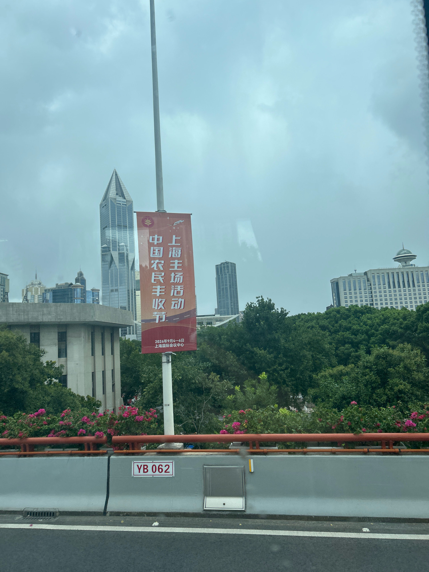 The image captures a street-level view of a city, likely Shanghai, China, under an overcast sky. In the foreground, a highway barrier is visible with a license plate reading "YB 062" mounted on it. Beyond the barrier, a vibrant display of pink flowers is seen, hinting at a park or landscaped area.
Dominating the middle ground is a tall, thin banner displaying Chinese characters in red on an orange background. The banner appears to announce an event, with smaller text at the bottom indicating "2026年9月4日—6日 上海国际会议中心" (September 4-6, 2026, Shanghai International Convention Center).
In the background, the cityscape is characterized by modern architecture. A distinctive, sharp-angled skyscraper stands prominently, while other buildings of varying heights and designs are scattered across the horizon. Lush green trees are interspersed throughout the urban landscape, softening the concrete jungle. The overall atmosphere is somewhat muted due to the cloudy weather.