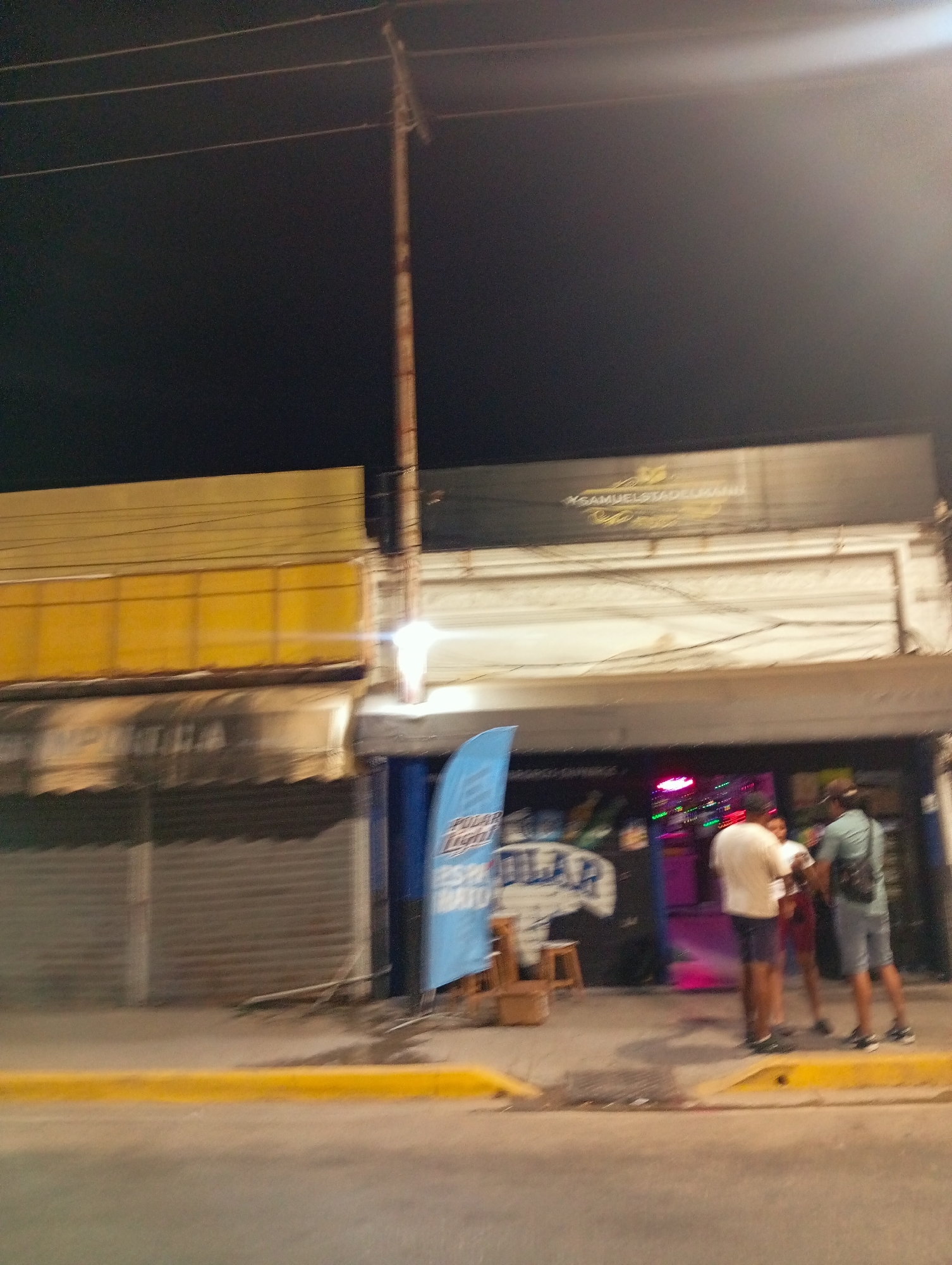 The image depicts a street scene at night in Juan Griego, Venezuela. Several people are standing outside a bar or establishment with brightly colored lights. There's a vertical banner in front of the building. A utility pole is visible. A store with metal roll up shutters is on the left. There is yellow curb.