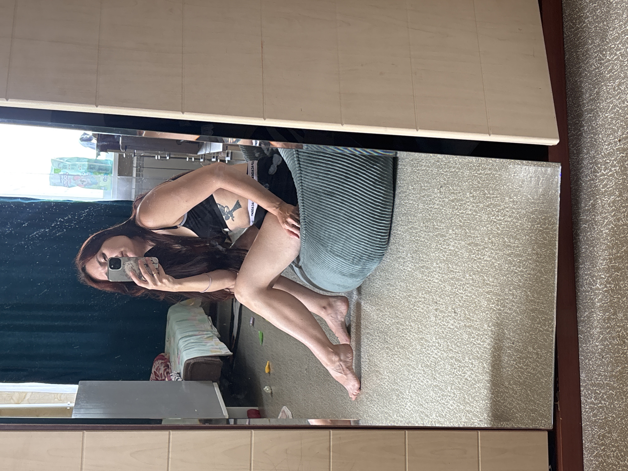 A young woman with long, dark hair is taking a selfie in a mirror. She is wearing a black sports bra and black shorts. She is sitting on a large, teal-colored beanbag chair. In the background, a bed with a light blue and white patterned comforter is visible, as well as a white closet with wooden doors. The room appears to be a bedroom, likely in an apartment in Naberezhnye Chelny, Russia.