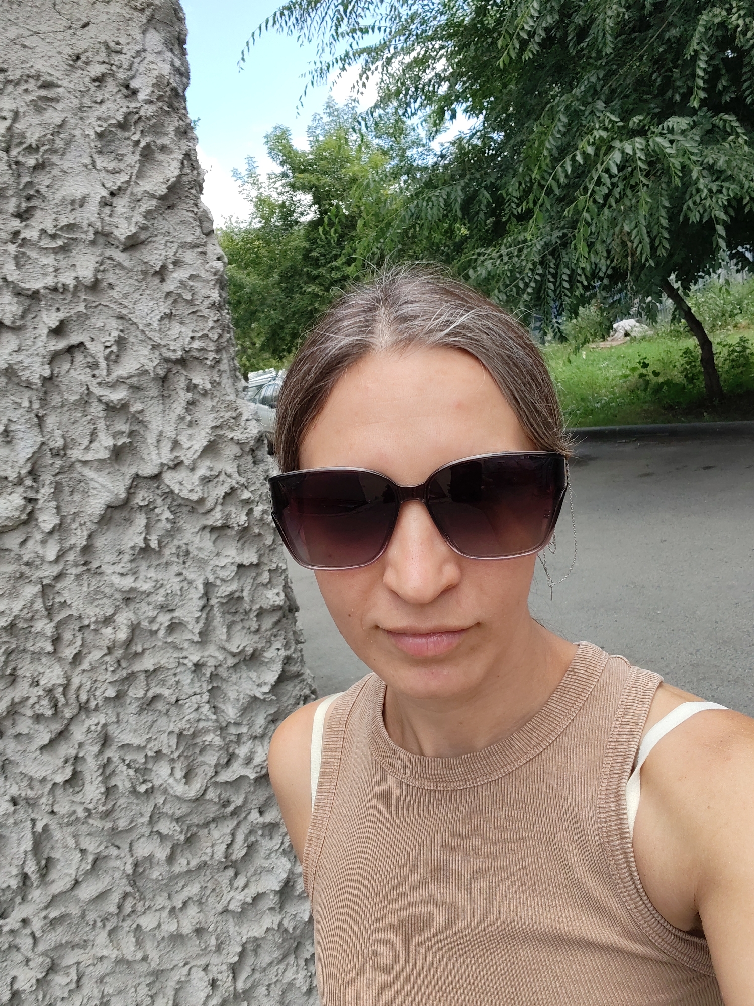 A woman is taking a selfie outdoors. She is wearing sunglasses and a beige tank top. To her left is a textured gray pillar, and behind her are trees and a paved surface. The sky is visible above the trees.