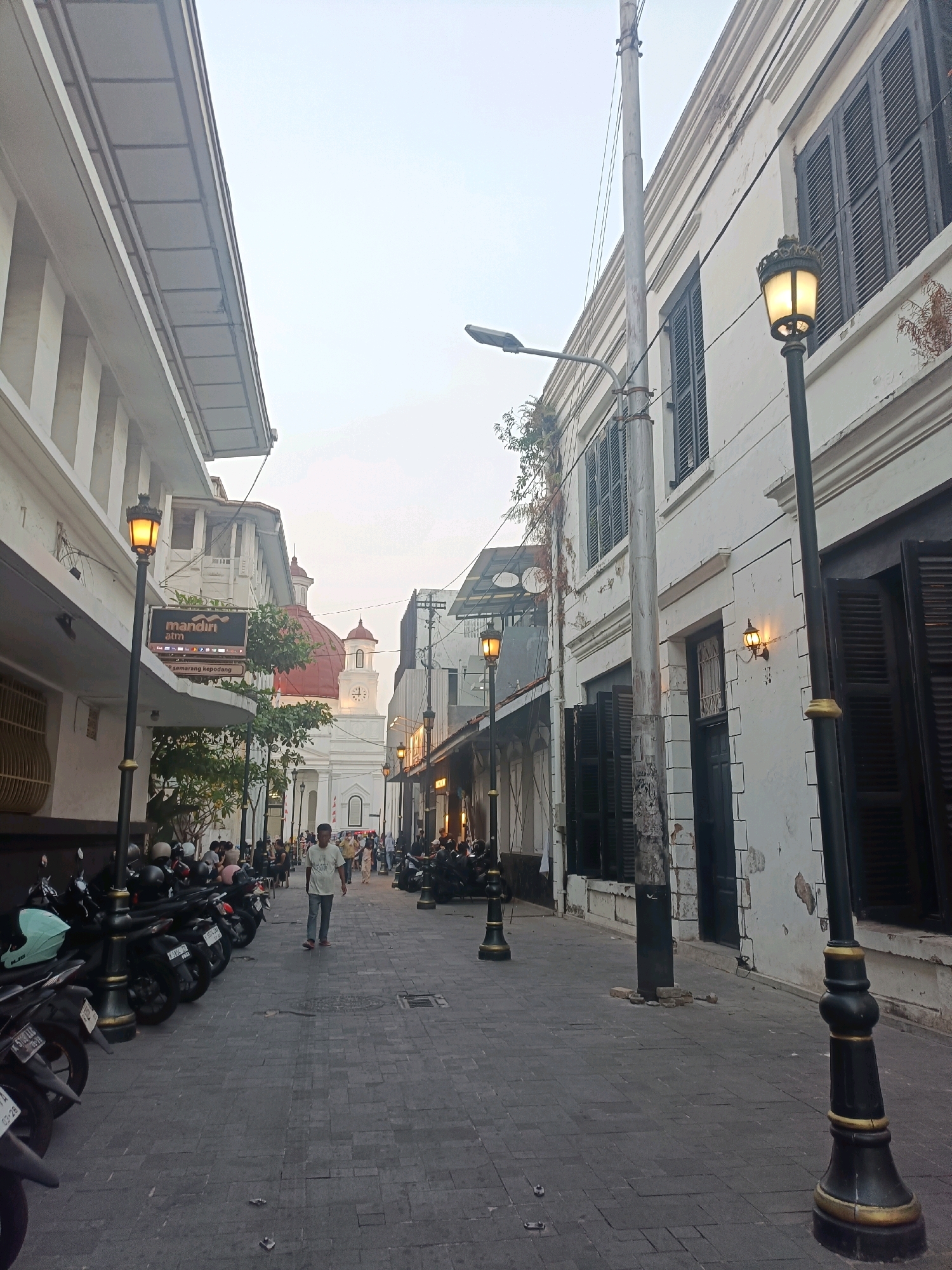 The image depicts a narrow street in the Kota Tua (Old Town) area of Semarang, Indonesia. The architecture is colonial, with white buildings on both sides of the street featuring dark shutters and ornate details. A prominent red-domed building with a clock tower is visible in the distance, suggesting it might be a church or historical landmark.
Several people are walking along the cobblestone street, some appearing to be tourists. The street is lined with parked motorcycles, typical for urban areas in Indonesia. Decorative street lamps, illuminated, are strategically placed along the pathway, adding to the ambiance of the historic district. The sky is overcast, indicating either early morning or late afternoon light. The overall impression is of a well-preserved historical urban environment.