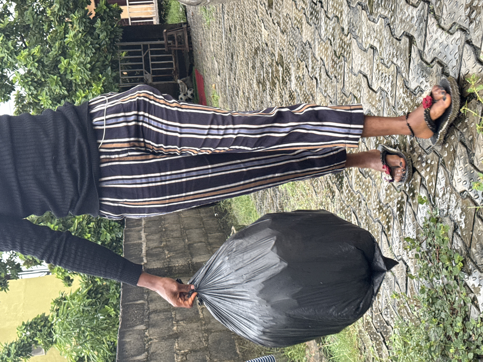 An individual stands on a patterned pavement. They are wearing striped pants, a black top, and sandals decorated with flowers. The individual is holding a large black trash bag in their right hand. The background includes trees, a building, and a brick wall. The location is Ado, Nigeria.