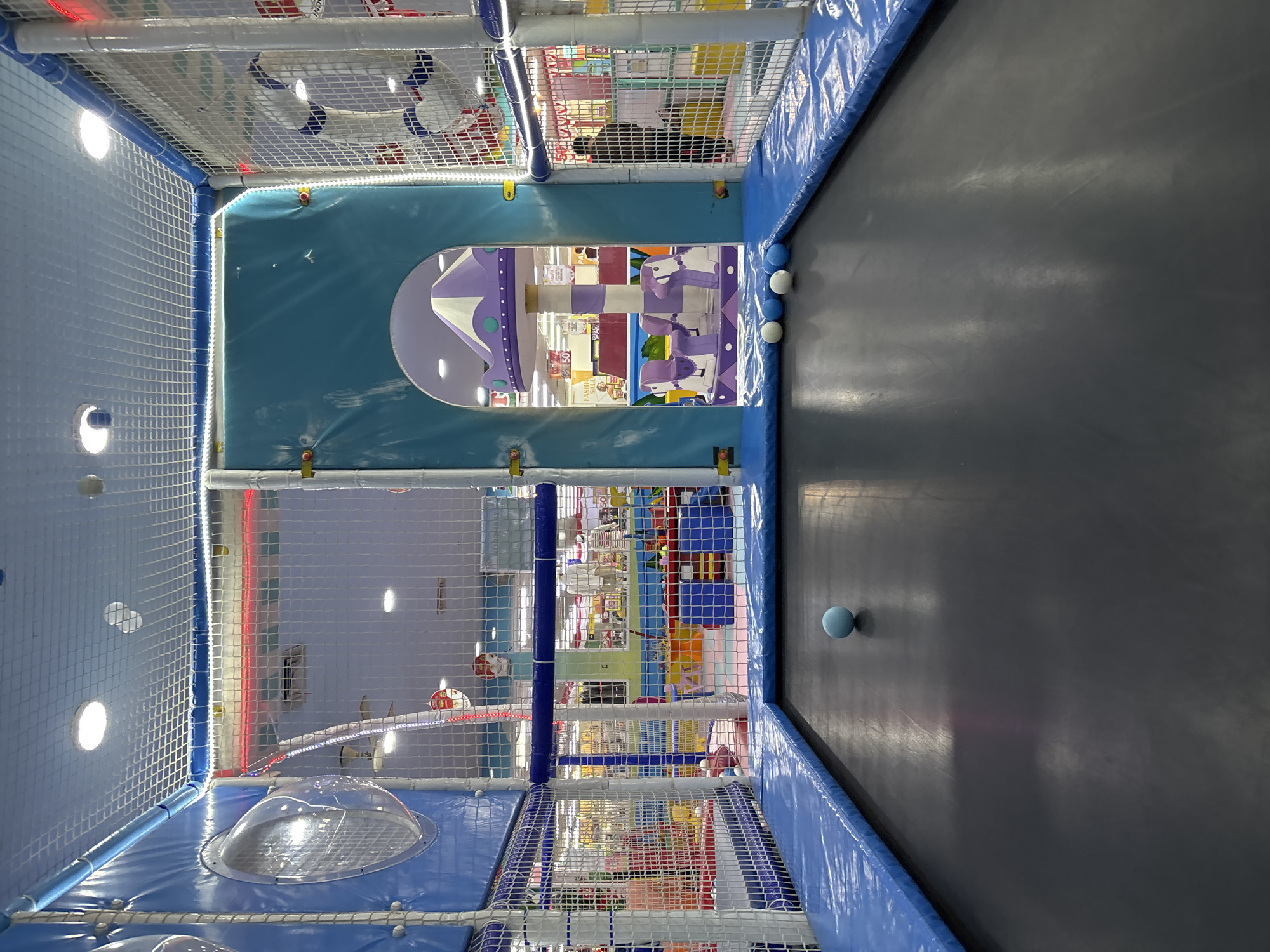 A wide-angle, eye-level shot captures the interior of an indoor children's play area, likely located in Liangjulang, Indonesia, based on the provided context. The foreground is dominated by a black trampoline surface with a blue padded border. A single light blue ball rests on the trampoline. Further back, a blue arched entryway opens into a colorful play structure. To the right of the entryway, the netting of the play area reveals glimpses of retail displays and shoppers beyond, suggesting this play area is situated within a larger commercial space. Multiple white and blue balls are visible near the entryway of the play structure. The environment is brightly lit with overhead lights and decorative LED strips.
