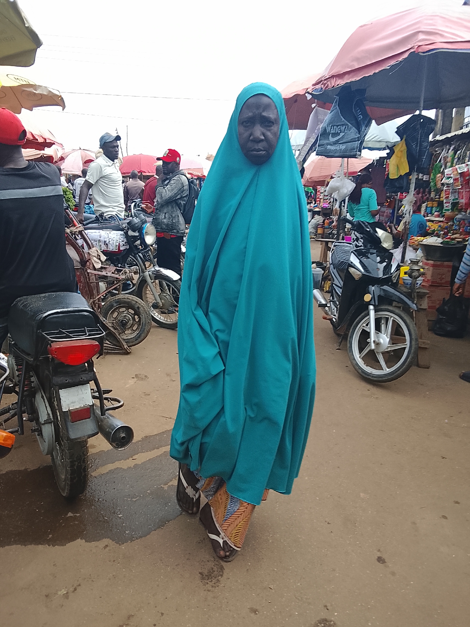 In Akwanga, Nigeria, a woman wearing a teal hijab and a patterned skirt stands facing forward in a busy outdoor market. She is partially obscured by her clothing. Behind her, several men are visible. One man on the left wears a black shirt and a red cap. Another man in the center wears a white shirt and a blue cap, and is standing near a motorcycle. To the right of him, another man wears a grey jacket and a red cap. Various stalls with umbrellas are set up in the background, displaying goods. Several motorcycles are parked throughout the scene, including one in the foreground on the left and another black motorcycle to the right of the woman. The ground is paved with dirt and there are some puddles, indicating recent rain.
