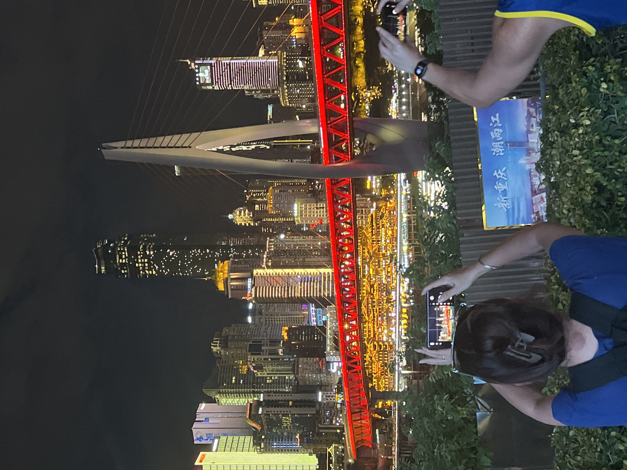The image shows a nighttime cityscape with numerous illuminated skyscrapers and a prominent red bridge spanning a body of water. In the foreground, two individuals are visible. The person on the left, seen from behind, is holding a smartphone to take a photograph of the cityscape. Their dark hair is styled with a clip. The person on the right, also seen from behind and from the waist up, has their right arm raised, potentially to take a photo or wave. They are wearing a blue and yellow athletic top and a smartwatch on their left wrist. Between the two individuals is a sign with Chinese characters and a depiction of a city skyline. The background is dominated by the dense urban architecture, characterized by a multitude of brightly lit buildings of varying heights and designs, and the illuminated red bridge structure. The overall scene suggests a tourist or visitor documenting the vibrant night view of a modern city.