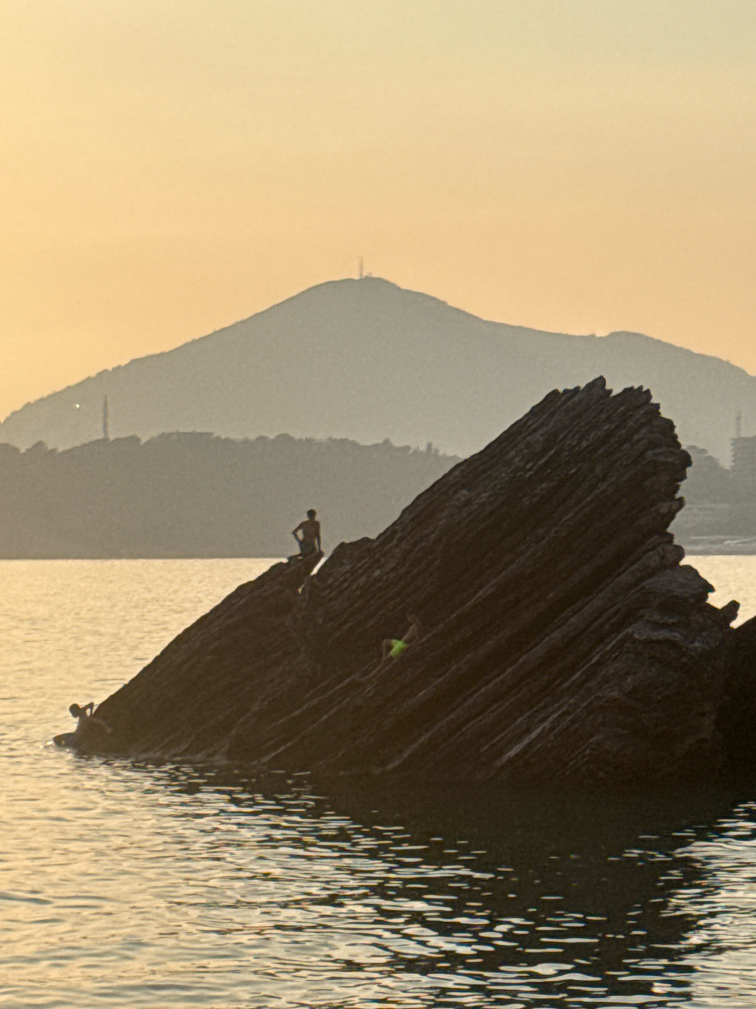 The image depicts a tranquil sunset scene at a rocky shoreline, likely in Kamenovo, Montenegro. In the foreground, a large, dark, jagged rock formation juts out from the water, its textured surface catching the warm, golden light of the setting sun. Several individuals are visible interacting with the environment. On the far left, a person is partially submerged in the water, appearing to be either swimming or wading. Further to the right, on the prominent rock, another person sits near the edge, silhouetted against the sky. A third individual is visible on the rock face, seemingly lying down. In the background, layers of hills and mountains recede into the distance, shrouded in a soft, hazy mist. A tall tower is visible atop the furthest mountain peak. The water is calm, reflecting the soft hues of the sky, with gentle ripples disturbing the surface. The overall atmosphere is serene and peaceful, capturing a moment of leisure as day transitions into evening.