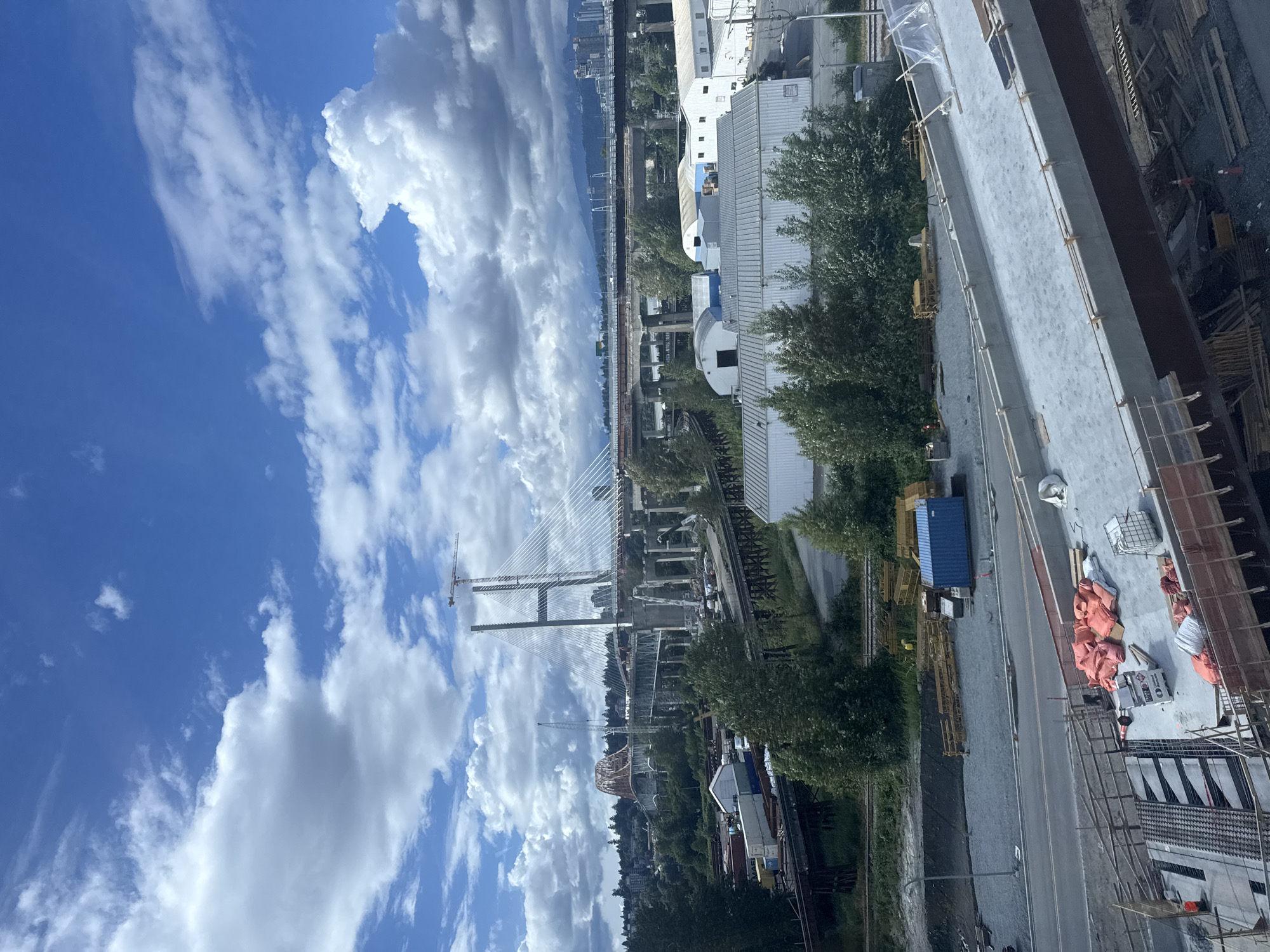 This image depicts a construction site in Surrey, Canada, focusing on a large bridge project. In the foreground, a section of a concrete bridge deck is under construction, with scaffolding, rebar, and orange safety barriers visible. Several piles of orange-wrapped materials, possibly construction supplies, are also present. Further in the background, a long span cable-stayed bridge dominates the scene, with its towers and numerous cables reaching towards the sky. A construction crane is positioned near one of the bridge towers, indicating ongoing work. Multiple older bridge structures are also visible, suggesting a complex, multi-stage project. Industrial buildings with metal roofs are situated below the bridges, and a body of water, likely a river, can be seen in the distance. Trees and greenery surround the construction area, and a cityscape with high-rise buildings and mountains forms the backdrop. The sky is blue with scattered white clouds. The scene appears to be during daylight hours.