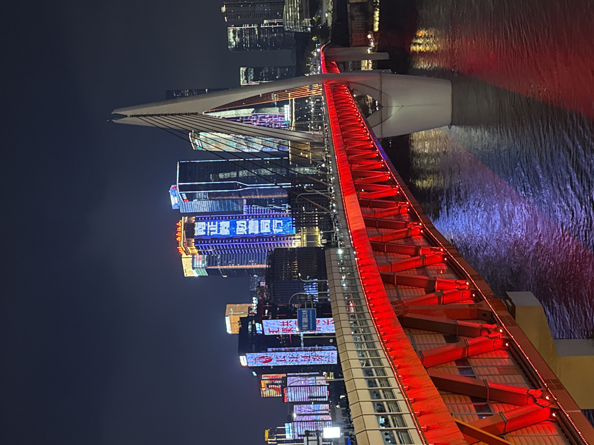 This is a night-time, medium-distance, eye-level shot of the Hongya Cave Bridge in Yuzhong District, China. The bridge spans a body of water, likely a river, which reflects the colorful lights of the city and the bridge itself. The bridge is illuminated with vibrant red lights along its structure and walkway. In the background, a dense collection of illuminated skyscrapers forms a city skyline, with various building facades displaying colorful LED signs in Chinese characters. The sky is dark, indicating nighttime. There are no discernible people in the foreground, but a few figures can be seen on the illuminated walkway of the bridge in the mid-ground. The overall scene is a vibrant display of urban nighttime illumination and architecture.