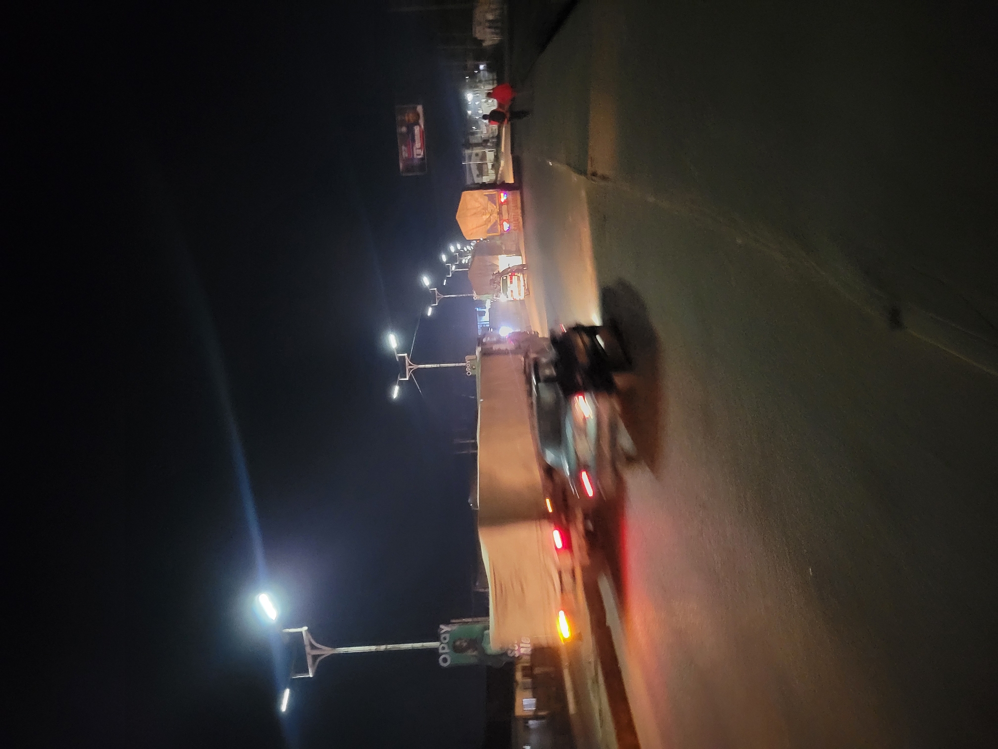 At night in an urban environment, a road is illuminated by streetlights and vehicle headlights. A semi-trailer truck is positioned on the road, its cargo covered with a light-colored tarp. Adjacent to the truck, a motorcycle is in motion. Farther down the road, a group of people, one of whom is wearing a red garment, are standing near another truck. A billboard with human faces is visible on the roadside. A sign with the text "Opay" is seen near a streetlight.