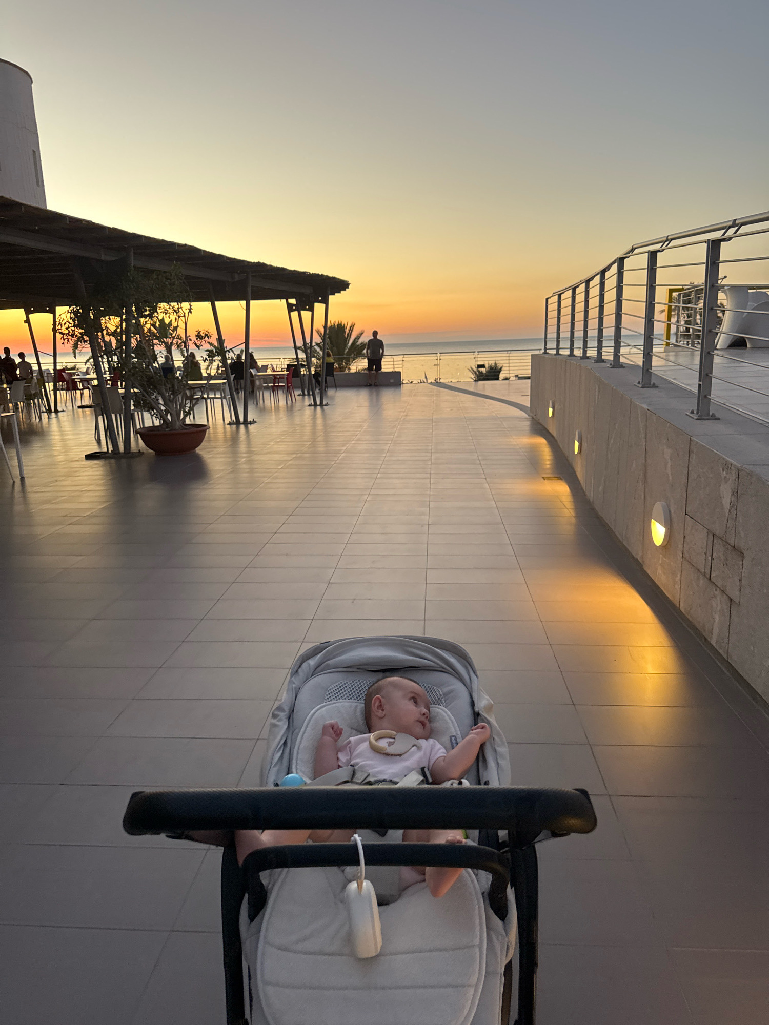 A wide-angle shot captures a sunset scene from the perspective of someone pushing a stroller. In the foreground, a baby is lying in the stroller, looking to the left. The baby is wearing a pink outfit and has a wooden toy attached to the stroller. The stroller itself is a light gray color and appears to be made of fabric.
The background reveals a tiled walkway with several outdoor tables and chairs, suggesting a dining area or a public space. Some people are visible in the distance, seated at tables or standing. A covered patio area with exposed wooden beams extends from the left side of the frame. Beyond the patio, the ocean is visible, with a strip of orange and yellow light indicating the setting sun.
To the right, a curved wall made of light-colored stone lines the walkway. Small, circular lights are embedded in the wall, casting a warm glow onto the tiled ground. A metal railing runs along the top of the wall, overlooking the ocean. In the far distance, a single figure stands silhouetted against the sunset. The overall lighting suggests late afternoon or early evening.
The image was taken in Isola delle Femmine, Italy, on the terrace of a restaurant or a similar establishment, likely overlooking the Mediterranean Sea.