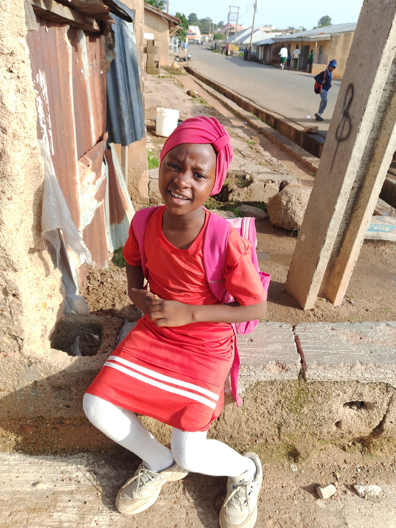 The media file shows a young girl in Jos, Nigeria, sitting on a ledge with a wall and corrugated metal to her left. She is wearing a red dress with white stripes, white socks, and beige sneakers. A pink backpack is slung over her shoulder. Her head is adorned with a pink headscarf. She has a joyful expression, smiling at the camera.
In the background, a street scene unfolds. Several people are walking along the road, and some buildings are visible. A concrete pillar stands to the right of the girl. The ground is uneven and appears to be made of dirt and broken concrete. The lighting suggests it is daytime, possibly late afternoon due to the shadows. The overall scene depicts a casual, outdoor environment in a Nigerian urban or semi-urban setting. There is no visible text in the image.