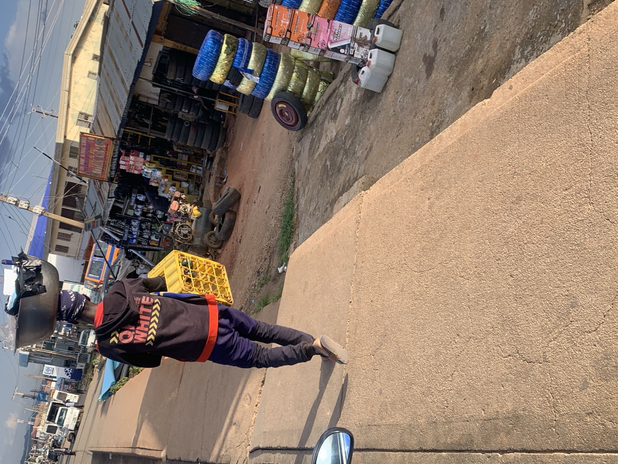 The image depicts a busy street scene in Jos, Nigeria, likely during the day. The perspective is from the ground, looking up and to the side.
In the foreground, a person is walking, carrying a large, round metal basin on their head, filled with various items covered by a plastic sheet. They are wearing a dark hooded sweatshirt with "ON WHITE" written in yellow block letters and red accents on the sleeves. They also have on dark pants and light-colored shoes. Beside them, a yellow plastic crate is visible.
The background reveals a roadside market with a row of stalls. One stall prominently displays stacks of tires, some of which are colored blue and yellow. Other stalls show an assortment of goods, including what appear to be boxes of footwear and various other items. Several vehicles, including trucks and a motorcycle, are also visible in the distance, suggesting an active commercial area.
The ground appears to be a mix of dirt and concrete. The sky is partly cloudy. The overall impression is one of a typical, bustling marketplace in Nigeria, with a focus on the person carrying goods on their head.
