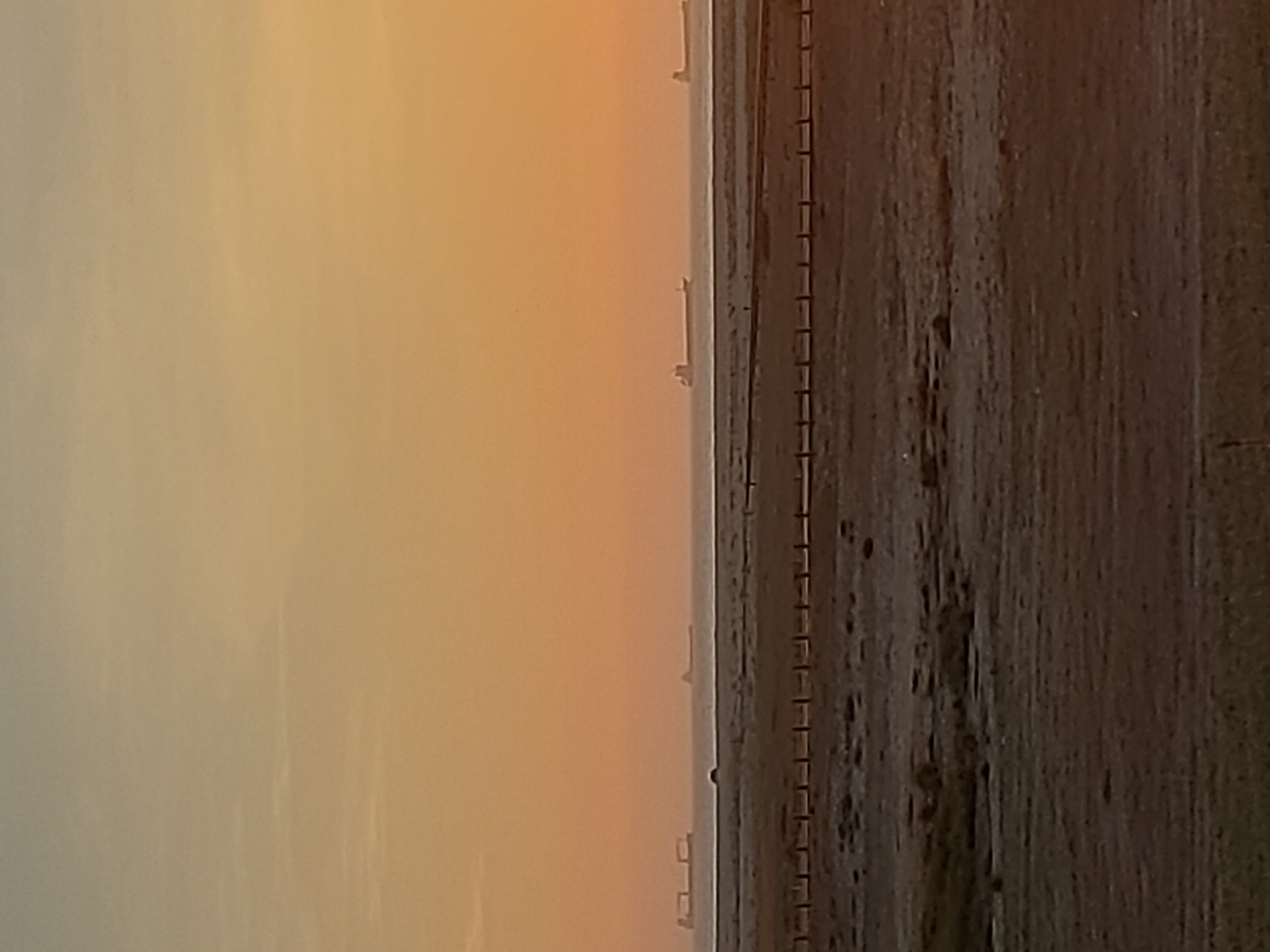 The image depicts a hazy, orange-tinged sky on the left, possibly suggesting either sunrise or sunset. Adjacent to the sky, a narrow strip of water is visible, possibly a river or the ocean. To the right of the water, a long, dark structure resembling a bridge or pier stretches into the distance. This structure features a series of regular, rectangular shapes along its side, suggesting a constructed element. Finally, the right side of the image is dominated by a dark, textured surface, possibly a road or pavement. The overall scene evokes a sense of a coastal or riverside setting, fitting for the location in the city of Figueira da Foz, Portugal.