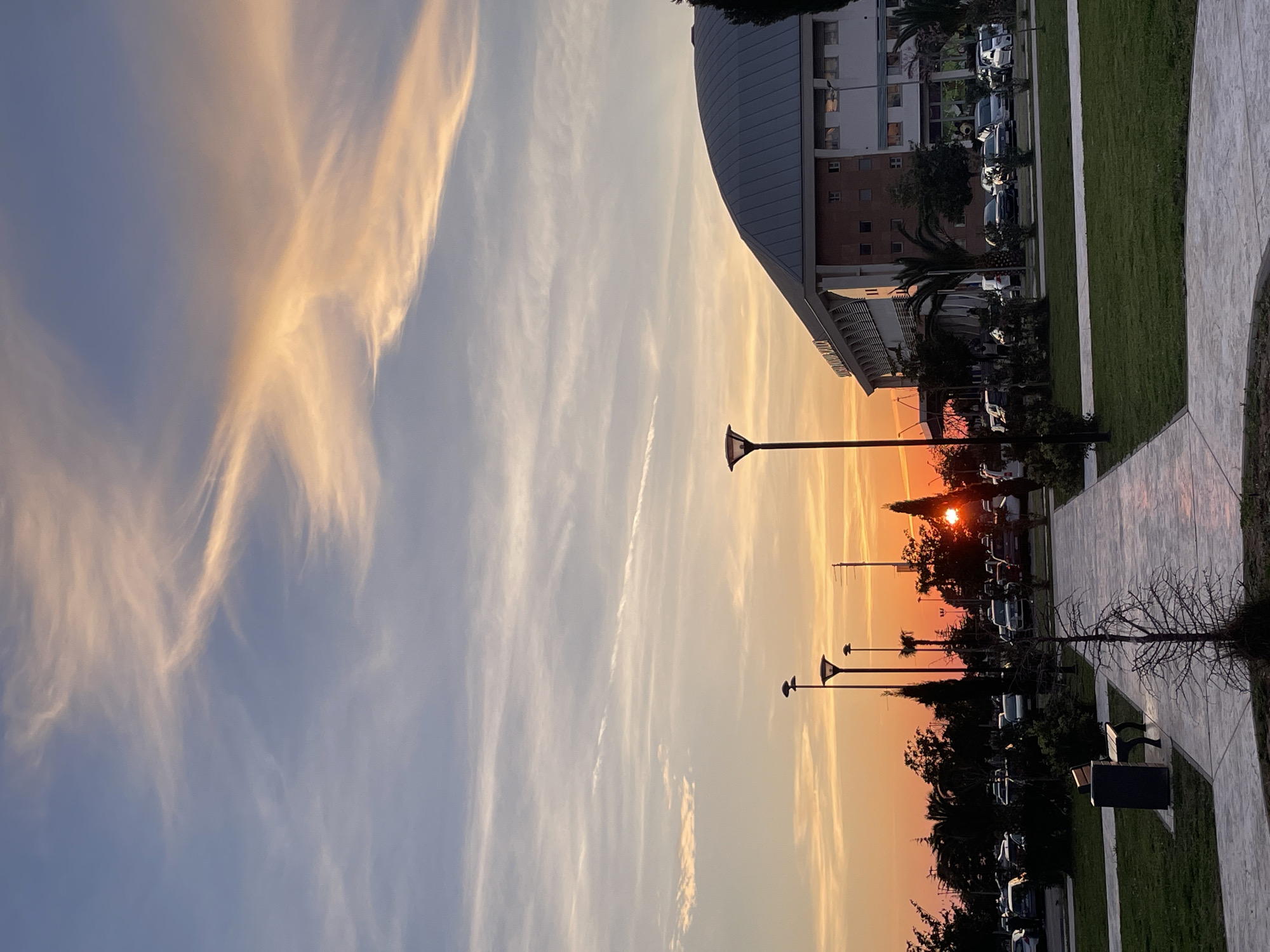 The image depicts an urban park scene at sunset in Bar, Montenegro, specifically featuring the Sportska Dvorana Topolica. The sky is marked by a clear horizon, where the sun, a bright orange orb, is partially obscured by distant trees and structures, casting a prominent solar flare. Above, cirrus and altocumulus clouds stretch across a gradient of colors from deep orange and pink near the horizon to pale blue overhead.
In the immediate foreground, a light-colored, paved pathway snakes through well-maintained green grass. To the left of the path, a dark park bench with a corresponding waste bin is positioned on the grass. A small, bare deciduous tree stands near the path's edge, casting a faint shadow.
The midground features several dark-colored streetlights with modern, tapered fixtures, standing tall along and around the pathway. A variety of trees, including cypress-like evergreens and palm trees, punctuate the landscape. Beyond these, a parking area is densely populated with numerous parked vehicles of various colors and models.
The background is dominated by the Sportska Dvorana Topolica, a large, multi-story building characterized by a distinct, expansive grey domed roof. Its facade is a combination of light-colored panels and brickwork, featuring multiple windows and a visible architectural detail resembling a sign or banner along its upper portion. Additional parked cars are visible adjacent to the building. The overall lighting is warm, indicative of late afternoon or early evening, with soft shadows and a general absence of direct human activity.