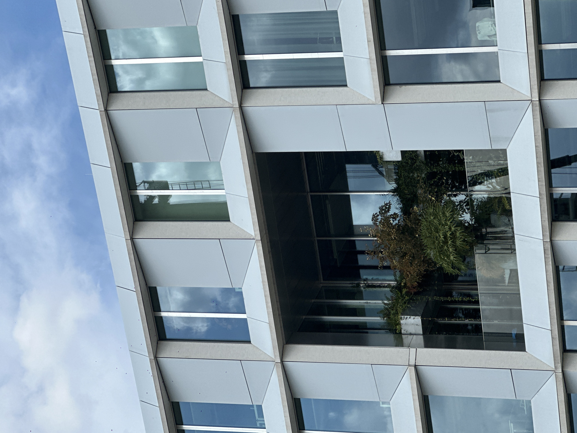 The image shows a modern building with a geometric facade, featuring large windows that reflect the sky and clouds. The building is composed of light gray panels with contrasting darker gray or metallic frames around the windows. The windows are arranged in a grid-like pattern, with some windows recessed and others flush with the facade.
In the central section of the building, there is an open space that reveals an interior balcony or terrace with lush greenery, including trees and hanging plants. A glass railing is visible on this balcony. The overall impression is one of contemporary architecture blending with nature.
The scene appears to be located in an urban environment, given the style of the building. There are no people visible in the image. The sky is partly cloudy, with a mix of blue sky and white clouds. The light suggests it is daytime.
The building's design is characterized by sharp angles and clean lines. The reflections in the windows are clear and detailed, showing the sky, clouds, and surrounding structures.
No specific landmarks or context suggest the city is Amsterdam, Netherlands.