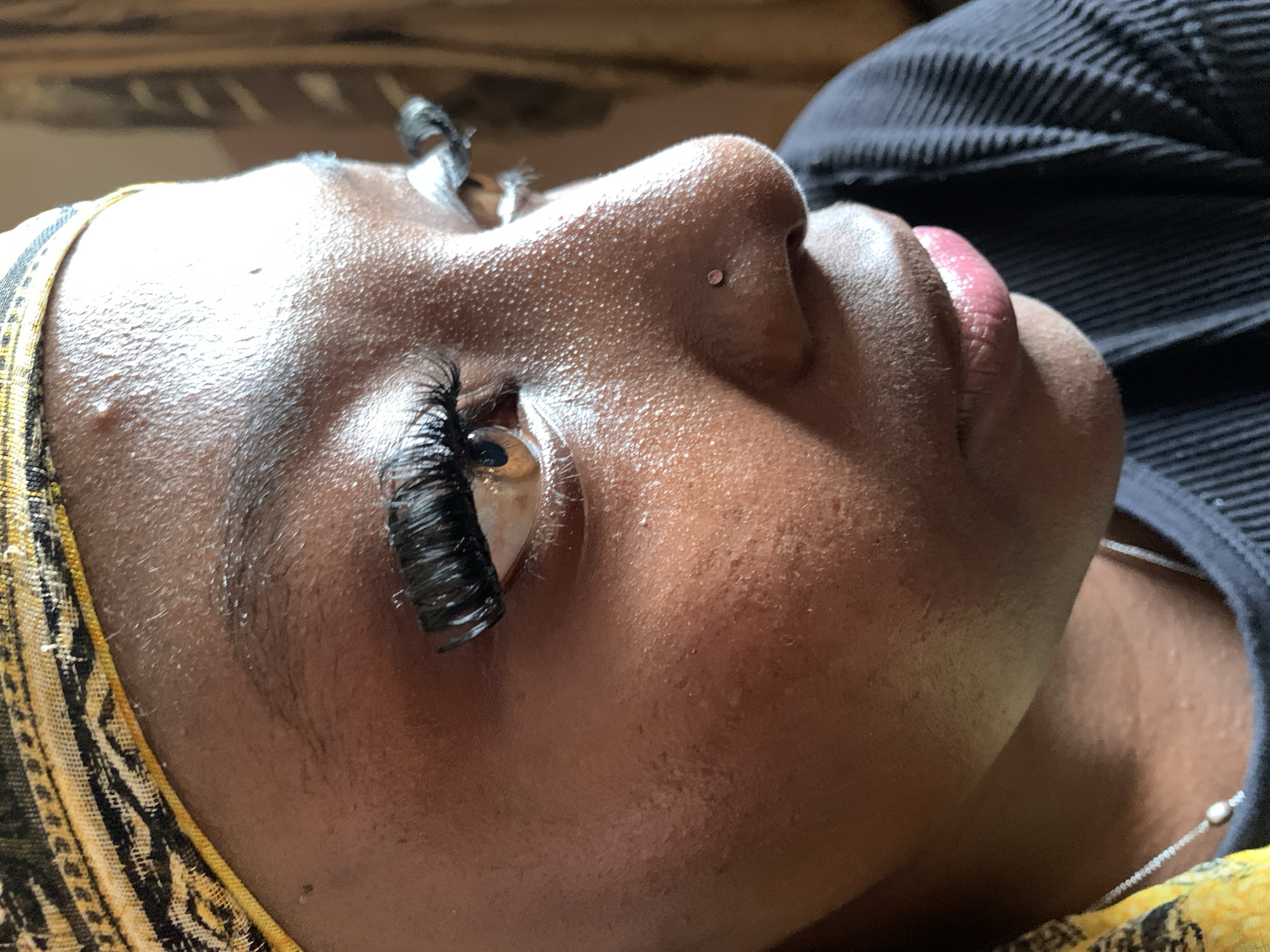 This is a close-up, eye-level, profile shot of a Black person lying down, with their head tilted upwards and to the left. The person is wearing dramatic, full eyelash extensions on their visible left eye, which is open and looking upwards. The person has a small, pink nose stud. Their skin has a textured appearance and appears to be slightly dewy or sweaty. They are wearing a yellow and black patterned headscarf that is partially visible on the left side of the frame, and a dark, ribbed garment on the right. The background is blurred, with horizontal lines suggesting wooden slats or blinds. The lighting appears to be natural and is casting shadows, suggesting it could be daytime. The overall impression is of someone undergoing or having just undergone a beauty treatment, possibly eyelash extensions, in a relaxed or reclined setting. The location context is Jos, Nigeria.