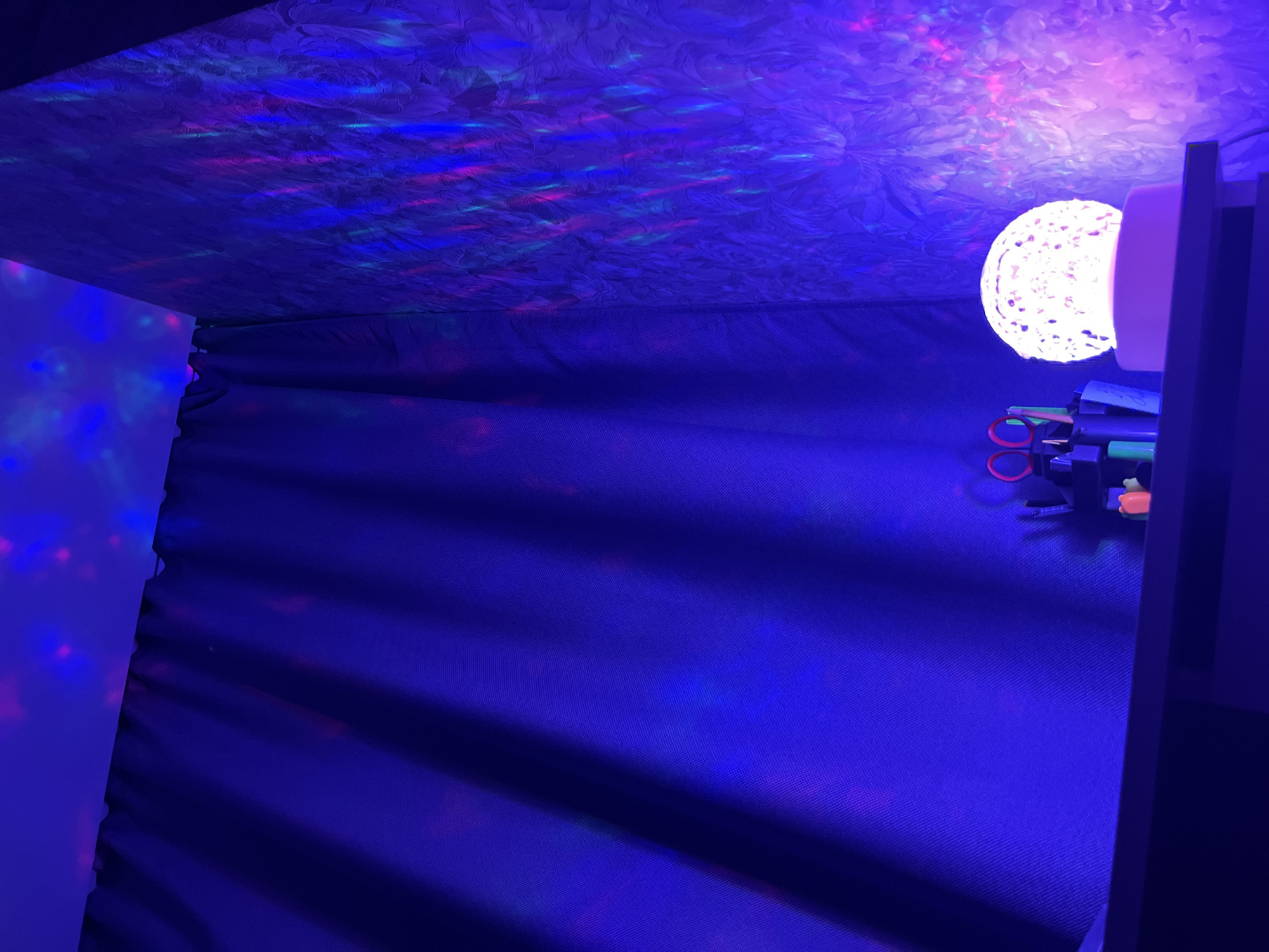 The image depicts a dark room illuminated by a projected light display of pink and blue dots on the ceiling and walls. A dark blue curtain hangs on the left side of the frame, obscuring part of the wall. On a dark shelf in the foreground, several items are visible: a light orb lamp, a set of scissors, pens, and some small colorful objects. The overall atmosphere is dimly lit and decorative. The image does not contain information to determine the location in the city of Krasnodar, Russia.