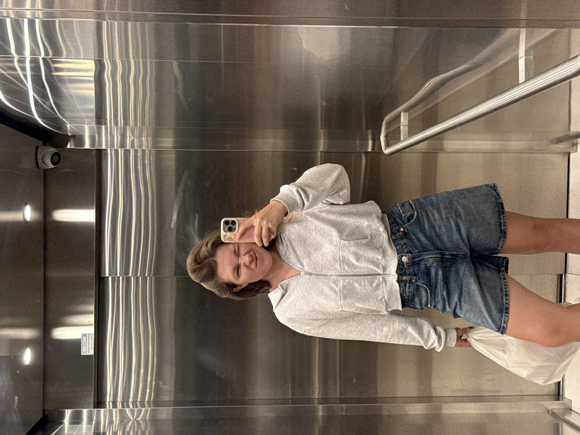 A woman is taking a selfie in an elevator. She is wearing a grey zip-up hoodie and denim shorts. She is holding a white plastic bag in her left hand. The elevator has stainless steel walls and a metal handrail. A security camera is visible on the ceiling. The location is not specified.