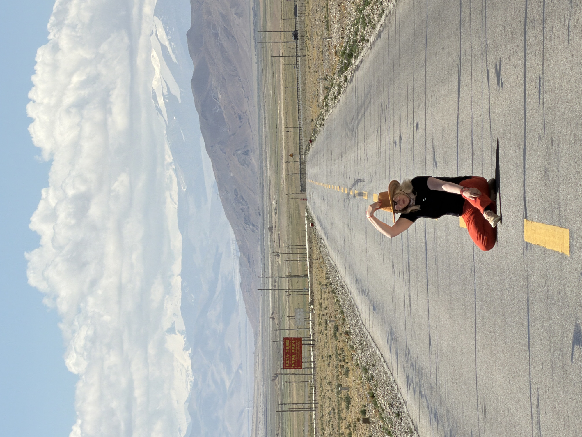 The image displays a vast, scenic landscape featuring a long, straight paved road cutting through an expansive plain. In the background, majestic mountains rise, some showcasing snow-capped peaks, under a bright blue sky dotted with large, fluffy white clouds. The setting suggests a high-altitude or remote region.
In the foreground, on the right side of the road, a person (likely a woman) is posing. She is wearing a black short-sleeved top, reddish-orange pants, and light-colored shoes. A light brown, wide-brimmed hat is perched on her head, and her hair appears blonde. She is in a crouching or kneeling position, looking towards the camera with a smile, her right hand near her hat.
The road features a yellow dashed line down its center. On both sides of the road, the ground is covered with dry, sparse vegetation, indicative of an arid or semi-arid environment. Utility poles or power lines line the left side of the road, extending into the distance.
A notable detail is a red rectangular sign with Chinese characters visible in the mid-ground on the left side of the road. Shadows cast by the person and other elements suggest it is late morning or early afternoon. The overall atmosphere is bright and sunny, conveying a sense of freedom and enjoyment in a wide-open, natural setting. The mountainous terrain and visible Chinese text are consistent with the provided location context of Mainpu township, China.