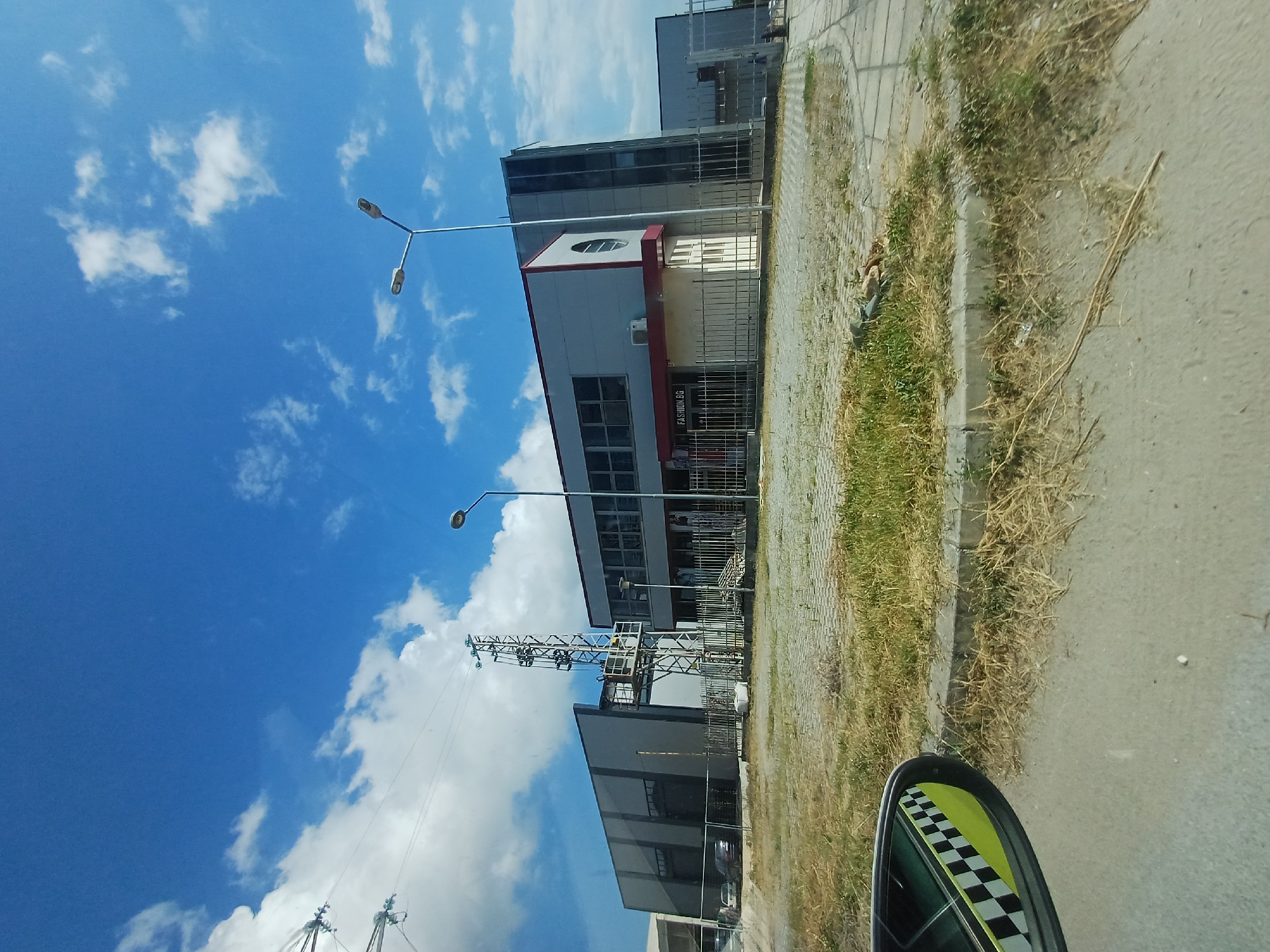 The image shows a street view in Aksakovo, Bulgaria, on a sunny day. The sky is a vibrant blue with scattered white clouds. In the foreground, a portion of a car's side mirror is visible, reflecting a black and white checkered pattern.
The main subjects of the image are buildings and infrastructure. There are several modern-looking buildings with glass windows and metallic facades. One building has a red-trimmed section with a circular window. Adjacent to the buildings, there's a paved sidewalk and a street. A streetlamp with multiple lights stands tall. In the middle ground, a metal utility structure, possibly a telecommunications tower, is visible. The area alongside the buildings and sidewalk is characterized by dry grass and weeds, suggesting a summer or dry season.
There is no clear activity or event taking place; it appears to be a static scene captured from a moving vehicle, indicated by the slightly tilted perspective and the presence of the car mirror. The time of day is likely daytime, given the bright sunlight and blue sky. The weather appears to be clear and warm. The location cues are limited to the text "FASHION.BG" visible on one of the buildings, which is likely a store name. No people are discernible in the image.