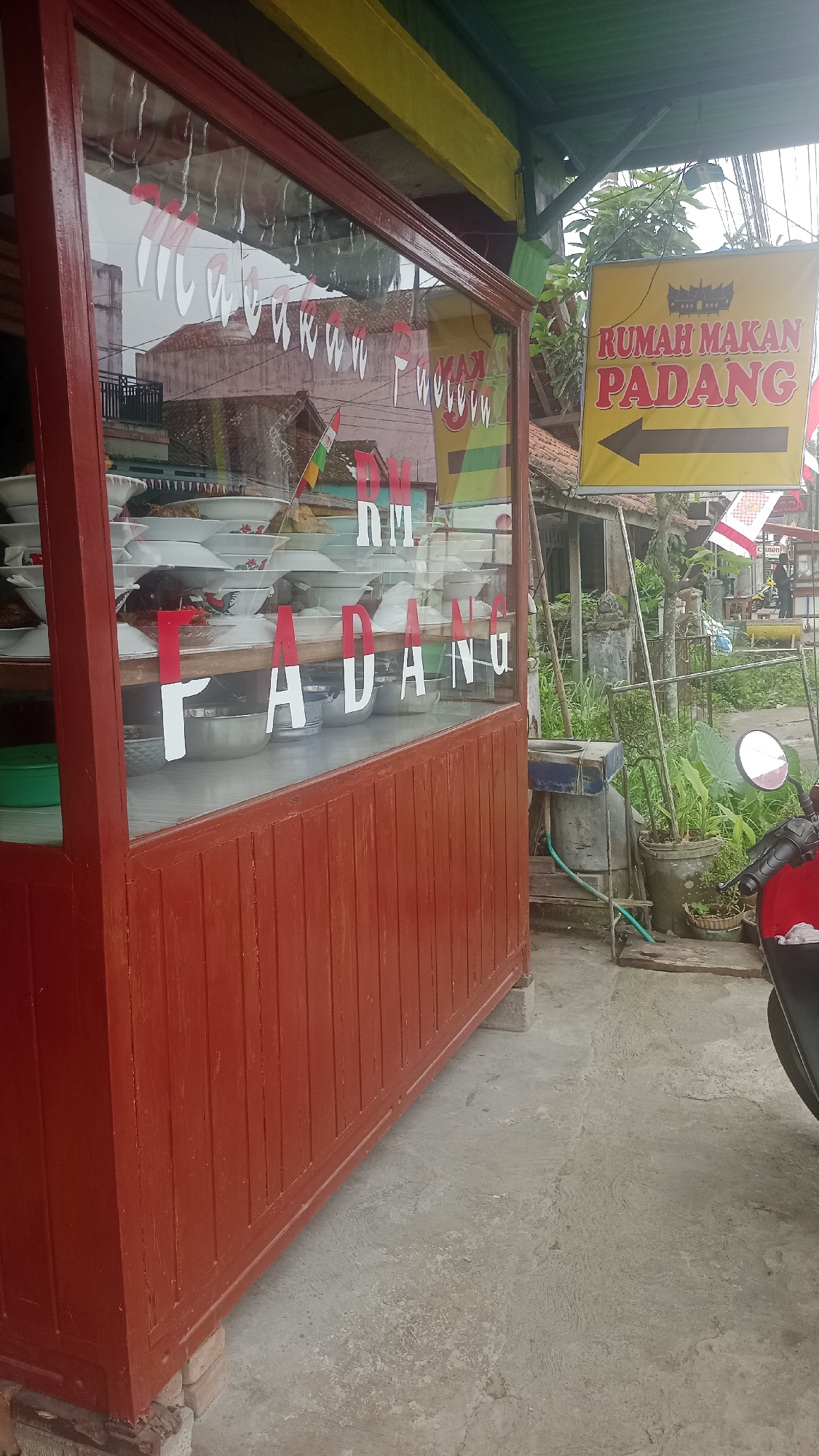 The image depicts the exterior of a restaurant in Pakemitan, Indonesia, featuring a prominent red wooden structure with glass windows. The windows display stacks of white bowls, likely containing food, and large white letters spelling out "PADANG," indicating it is a Padang restaurant. A yellow sign with red text, also reading "RUMAH MAKAN PADANG" and featuring an arrow pointing to the right, hangs to the right of the restaurant entrance. The scene is outdoors, with a concrete ground surface and some greenery visible in the background, including plants in pots and trees. A red motorcycle is parked on the right side of the frame. The overall impression is that of a small, local eatery in a somewhat casual, possibly rural or suburban setting.