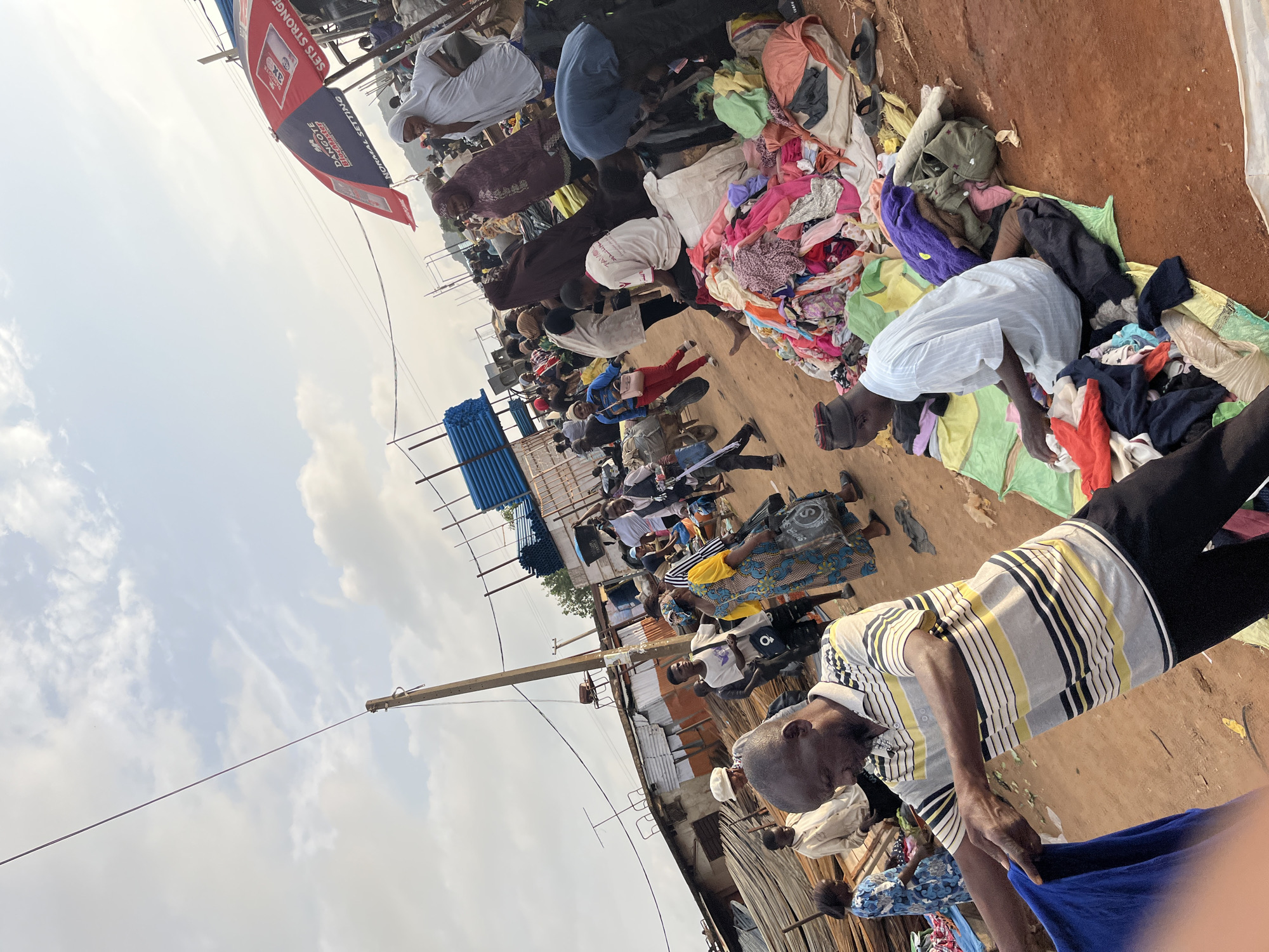 A daytime, outdoor, low-angle, full shot shows a bustling marketplace scene in Gura Top, Nigeria. The ground is red dirt, and the sky is cloudy and bright. In the foreground, a man in a striped polo shirt is holding up a blue garment, seemingly inspecting it. To his right, a pile of colorful clothes is spread out on a mat. A man in a white striped shirt sits on the mat amongst the clothes. Behind them, a diverse crowd of men and women, some in traditional attire, are browsing various stalls and piles of goods. Blue pipes are stacked on the left side of the image. A red and white umbrella, branded with "DANGOTE", is visible in the background. The overall impression is one of lively commerce and everyday activity in an open-air market.
