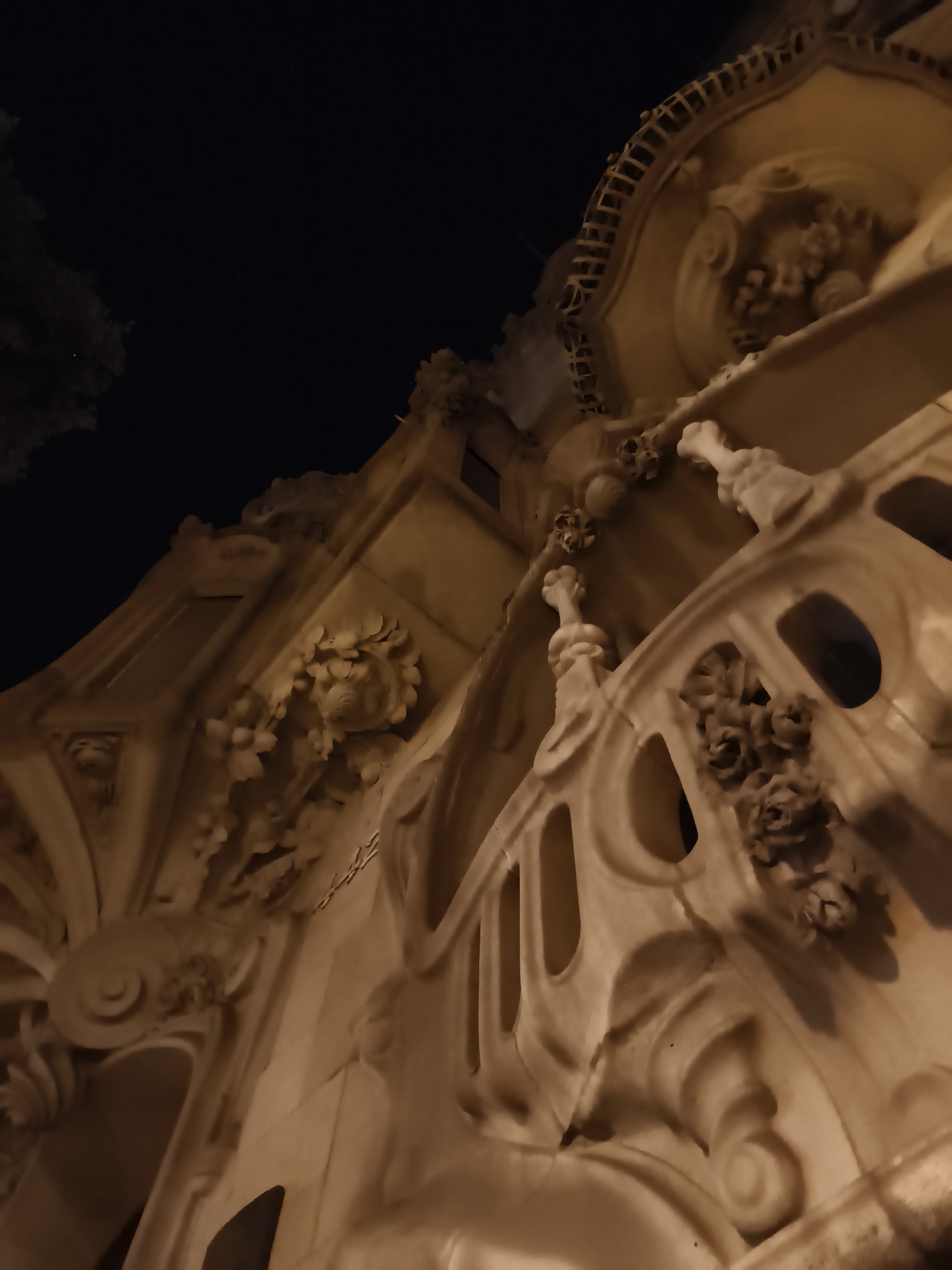 The image shows a low-angle, nighttime shot of the ornate facade of Park Güell in Barcelona, Spain. The lighting casts warm tones on the intricate stone carvings, highlighting floral motifs and architectural details like balustrades and decorative elements. The dark sky above provides a stark contrast to the illuminated building. There are no people visible in the image.