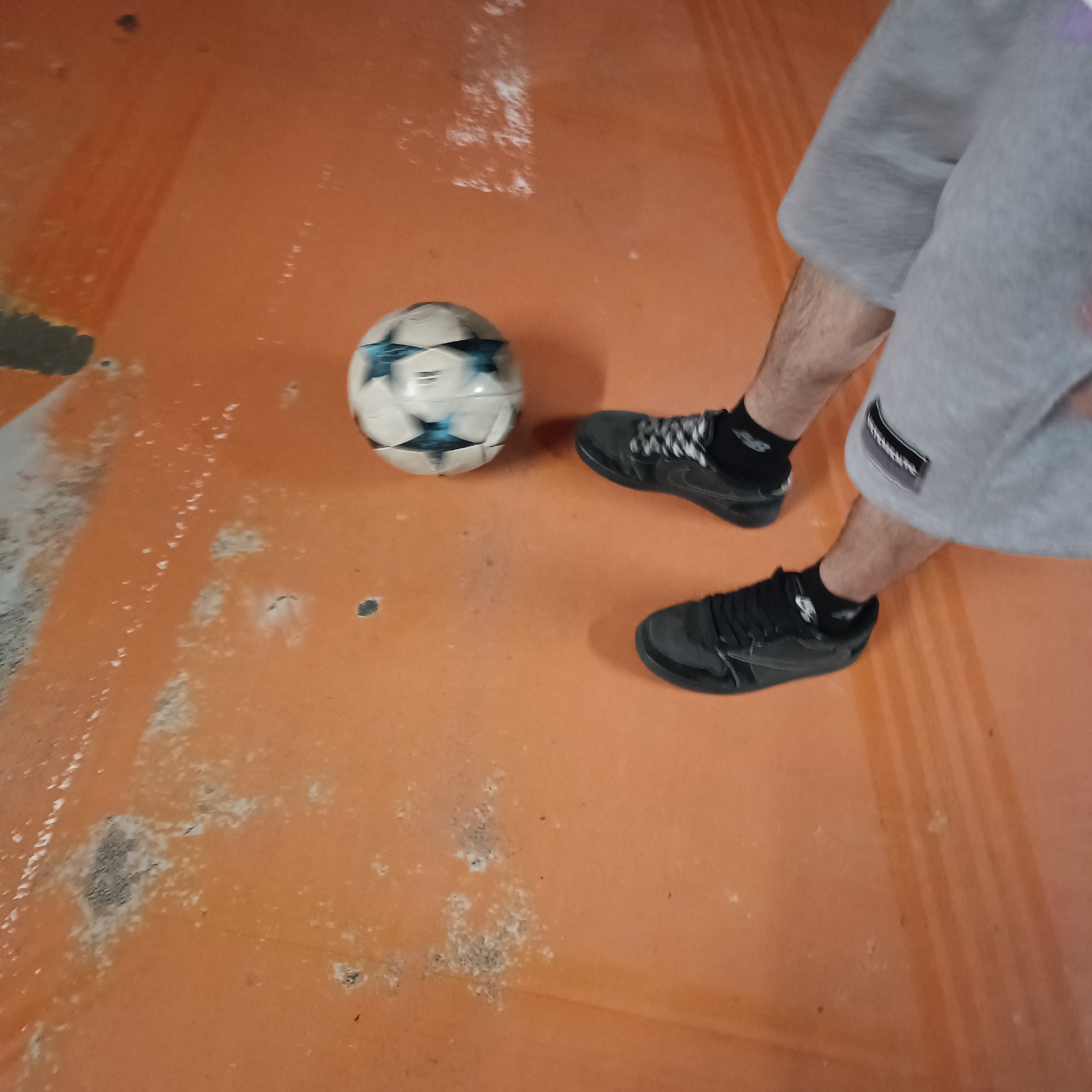 This close-up image shows the lower body of a person standing on an orange indoor court next to a soccer ball. The person is wearing grey shorts, black athletic socks, and black athletic shoes. The shoes feature black and white patterned laces; one shoe clearly displays a white swoosh logo, indicating it is likely a Nike brand. A black patch with white, unreadable text, possibly "Hampshire," is visible on the right thigh of the grey shorts.
A white and light blue/grey soccer ball, decorated with star-shaped patterns suggestive of a UEFA Champions League design, rests on the worn court. White text, likely brand details such as "adidas" and "Official Match Ball," is partially visible but unreadable on one of the ball's panels.
The court itself is a vibrant orange, showing considerable wear and tear, including scuff marks, lighter streaks, and darker patches of dirt or debris, indicating an active sports environment. The lighting is consistent with an indoor setting, with no direct sunlight visible, making the time of day indeterminate. The person's stance suggests they are either about to play, in the middle of playing, or have just finished a game of soccer or futsal. The location context provided is Sofia, Bulgaria.