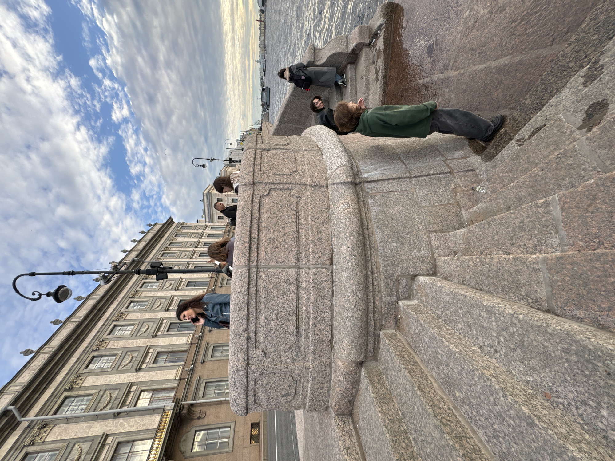 This is a medium shot photograph of a granite embankment and staircase along the Neva River in Saint Petersburg, Russia. A woman in a blue jacket is talking on her phone on top of the embankment, which is adorned with decorative molding. Several other people are visible on the embankment and the steps, including a young person in a green hoodie looking down at the water. The Neva River is wide and choppy, with a boat visible in the distance. The architecture of a grand, classical building is in the background, consistent with the historic St. Petersburg cityscape. The sky is partly cloudy with a mix of blue and white. The light suggests it is late afternoon or early evening.