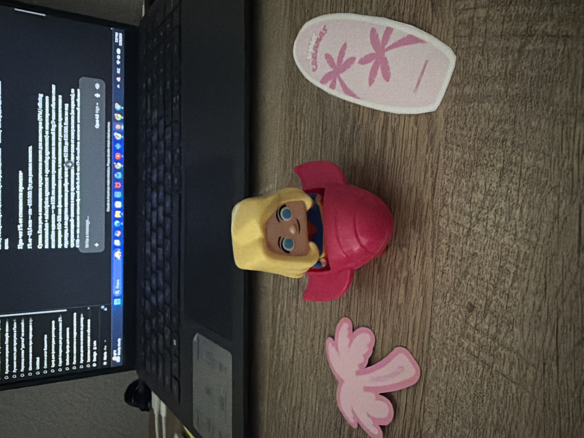 The image depicts a close-up overhead view of a wooden desk surface. In the foreground, from left to right, are three items: a pink, stylized palm frond cutout; a small, pink toy airplane with a blonde-haired, blue-eyed doll figure inside; and a pink flip-flop cutout with two pink palm tree graphics and the word "CABANAS" printed in pink. Behind these objects, a portion of a dark laptop is visible, open and displaying text on its screen. The overall scene suggests a casual or work-from-home environment. There is no discernible indication of the city of Orlando, United States of America, within the image itself.