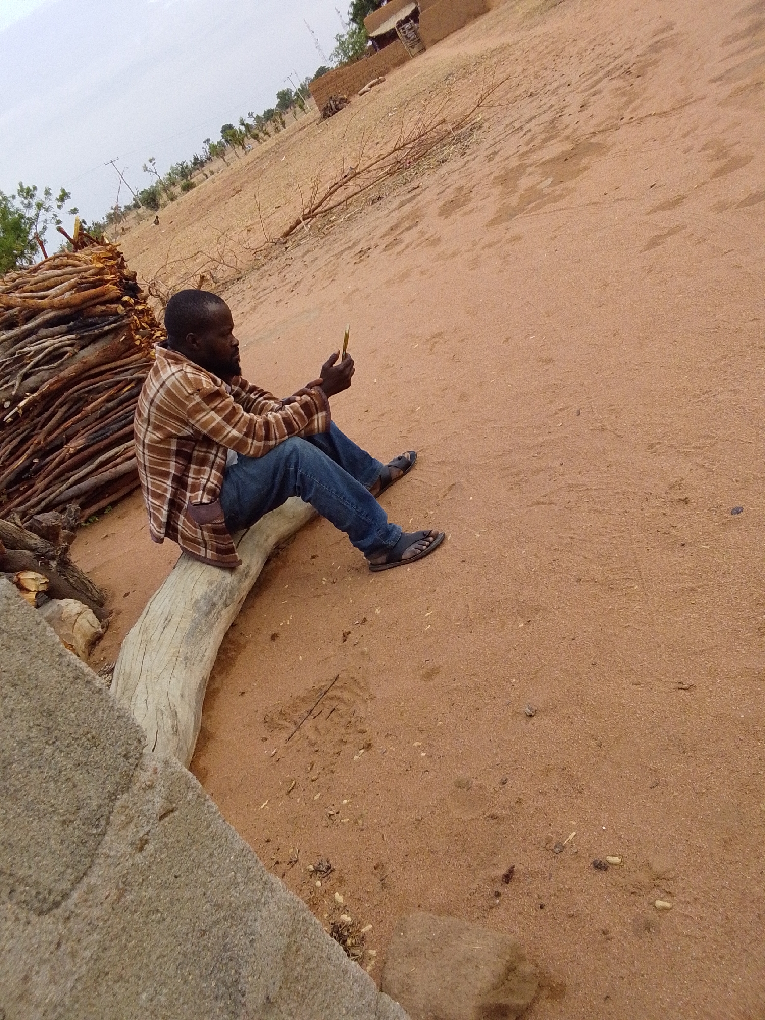 A man sits on a weathered log in a sandy environment. He wears a brown plaid shirt, blue jeans, and sandals and holds a small yellow object in his hands, appearing to examine it. Behind him is a pile of branches. The backdrop consists of a flat, sandy landscape leading to a few buildings in the distance under a cloudy sky. The location is Mulke Barikin Sayejo, Nigeria.