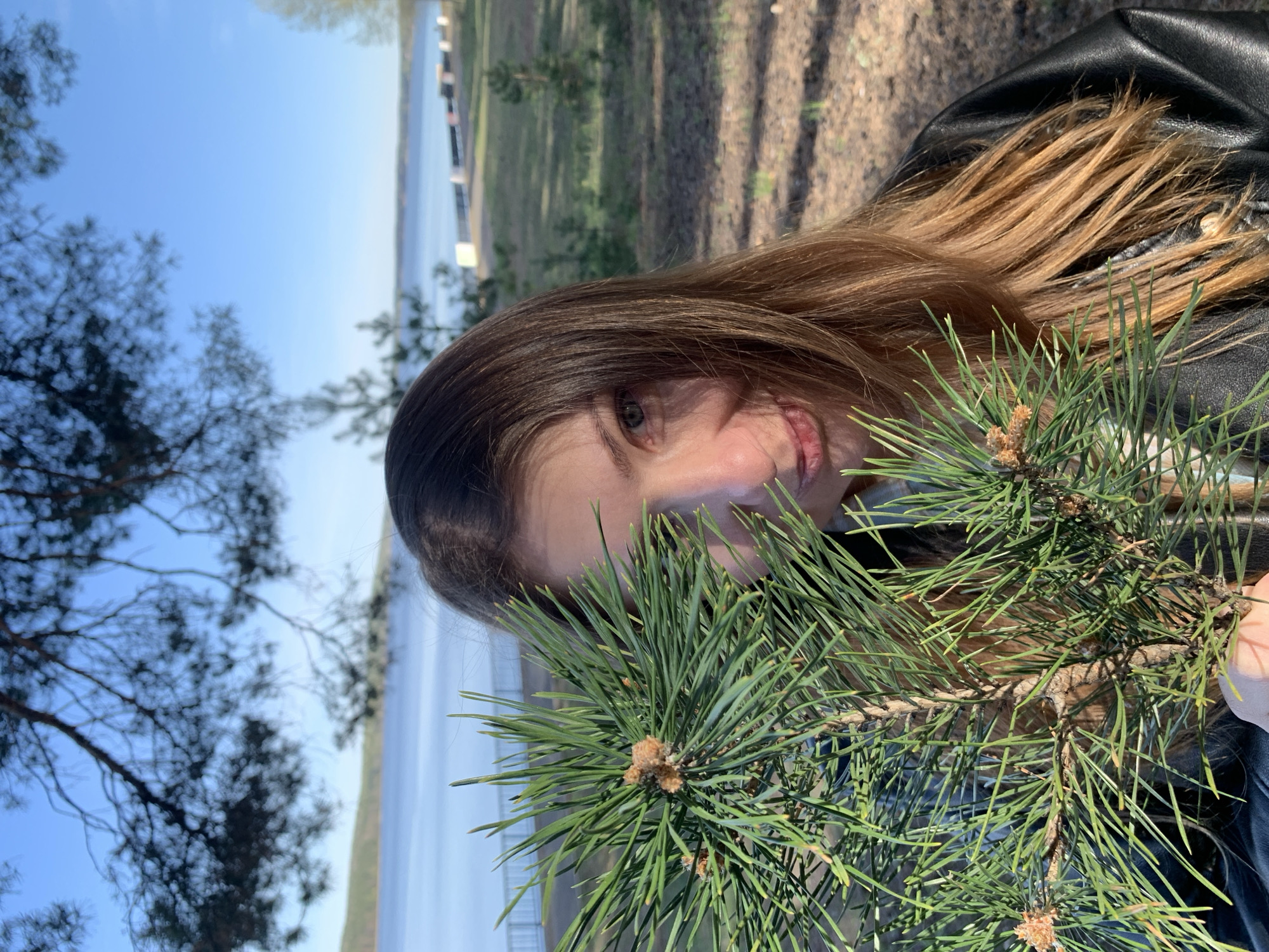 A young woman holds a pine branch in front of her face. She has medium-length brown hair and is wearing a black jacket. Behind her is a body of water with a visible shoreline and trees. The location is Chaikovsky, Russia.