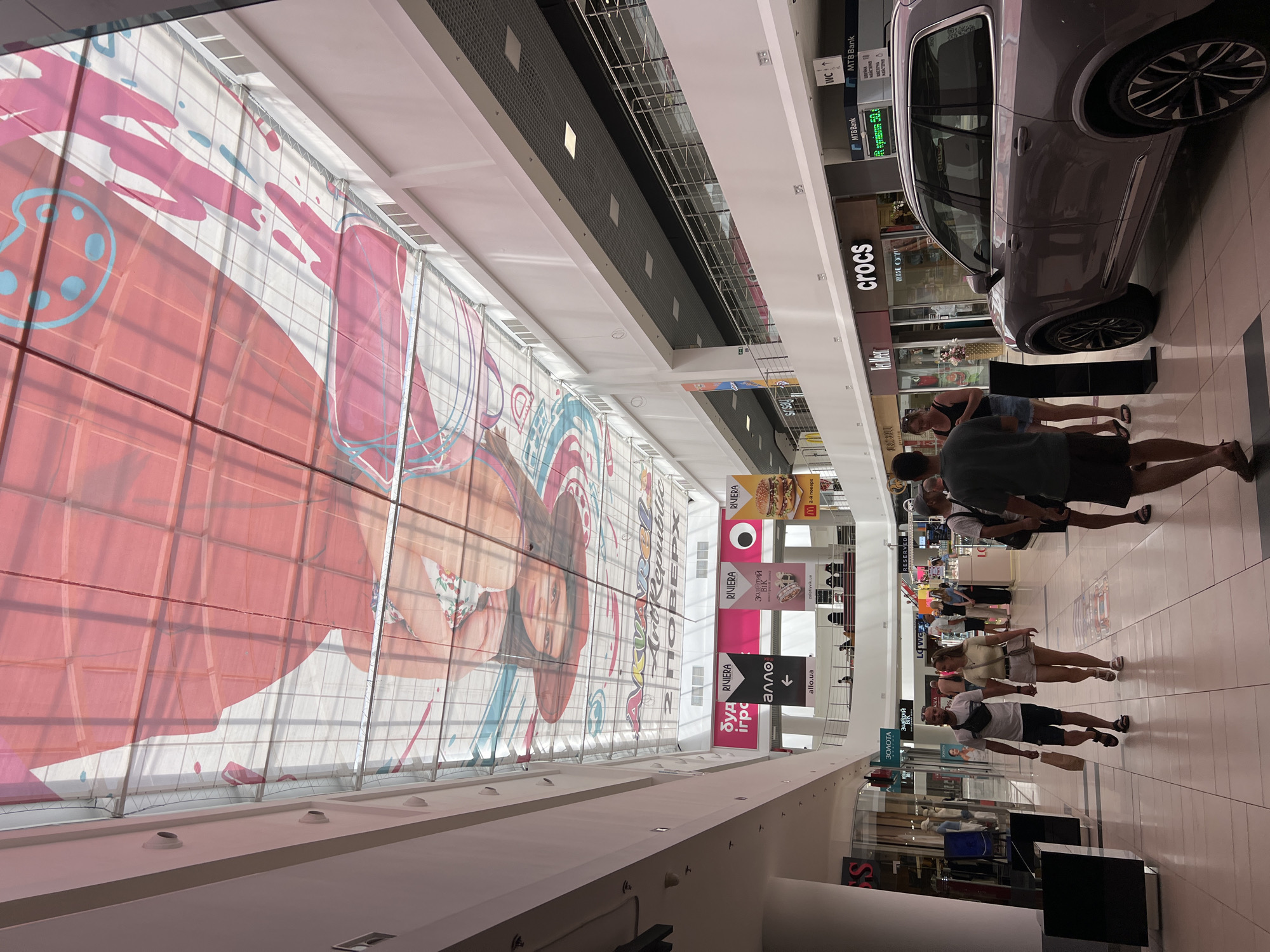 The image depicts the interior of a modern shopping mall. The ceiling is a large, translucent canopy adorned with a vibrant, abstract graphic featuring shades of pink, red, and blue, resembling paint splatters and a stylized illustration of a person. Natural light filters through this canopy, illuminating the spacious interior.
The mall features multiple levels with balconies and escalators visible. Storefronts line the walkways, with various brands such as "Riviera," "Allo," "Crocs," and "MTB Bank" being identifiable from their signage. One prominent advertisement displays a large image of a hamburger, suggesting a food establishment.
In the foreground, a couple is walking hand-in-hand down the main corridor. The man is wearing a white t-shirt, dark shorts, and a black fanny pack, carrying a paper shopping bag. The woman is in a light yellow t-shirt and white shorts. Other shoppers are dispersed throughout the mall, some walking and others browsing in the shops or resting on benches.
A light-colored SUV is parked on the right side of the frame, positioned near some of the storefronts. The floor is tiled, reflecting the ambient light. The overall atmosphere suggests a busy, commercial environment. The location is identified as the city of Lysychansk, Ukraine.