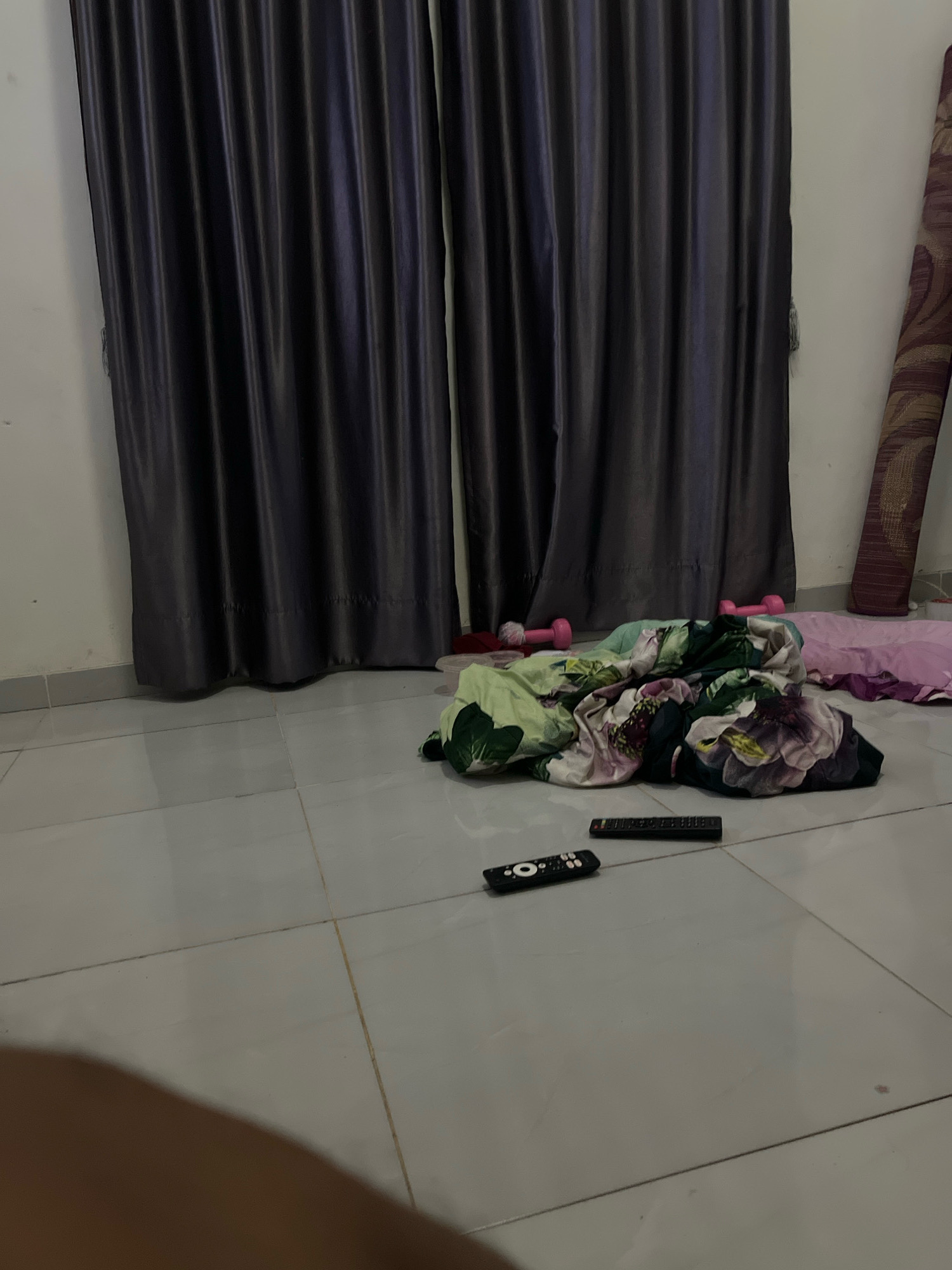 In an indoor residential setting, likely a bedroom, located in Kebumen, Indonesia, a scene unfolds with several objects scattered on a tiled floor. Dominating the background are dark, floor-to-ceiling curtains, drawn closed, suggesting either privacy or blocking out light. To the right, partially visible, is a rolled-up mat, possibly a sleeping mat or a rug.
In the mid-ground, a pile of bedding, featuring a floral pattern with prominent pink and green hues, lies crumpled on the floor. Adjacent to this bedding, two pink dumbbells are visible, hinting at a possible exercise area or personal fitness equipment. Nearby, a pink pillow is also present.
In the foreground, two remote controls rest on the tiled floor. One is black with a circular button at the top, and the other is a longer, black remote with numerous buttons. The floor is made of large, light gray square tiles, with visible grout lines. A shadowed area in the bottom left corner suggests the presence of a person partially obscuring the camera's view. The overall impression is a casual, lived-in space with items not meticulously stored.