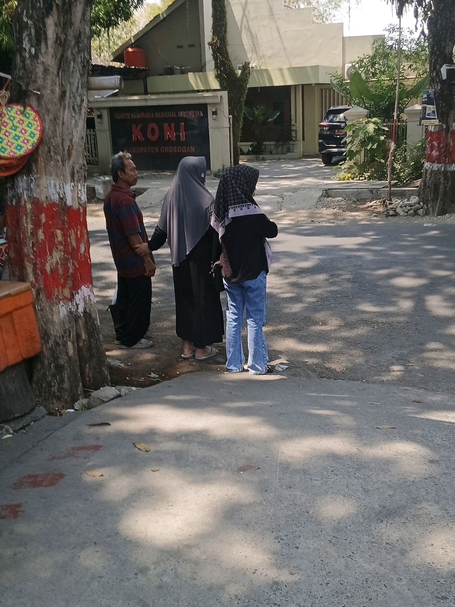 The image captures a street scene in front of the KONI (Komite Olahraga Nasional Indonesia) building in Purwodadi, Indonesia, on a sunny day with dappled sunlight casting shadows. In the foreground, a man wearing a striped polo shirt and dark pants stands on the sidewalk, facing away from the camera. Next to him, a woman in a dark hijab and black dress is also facing away, holding the man's arm. Further to the right, another person in a hijab and blue bell-bottom jeans stands with their back to the viewer. A black SUV is parked in the driveway of the building. The building itself has a light-colored facade with the "KONI" logo prominently displayed in red. The street is paved, and there are trees and vegetation lining the sidewalk and surrounding the building. An orange crate is visible on the left side of the image, next to a tree trunk with red and white markings.
