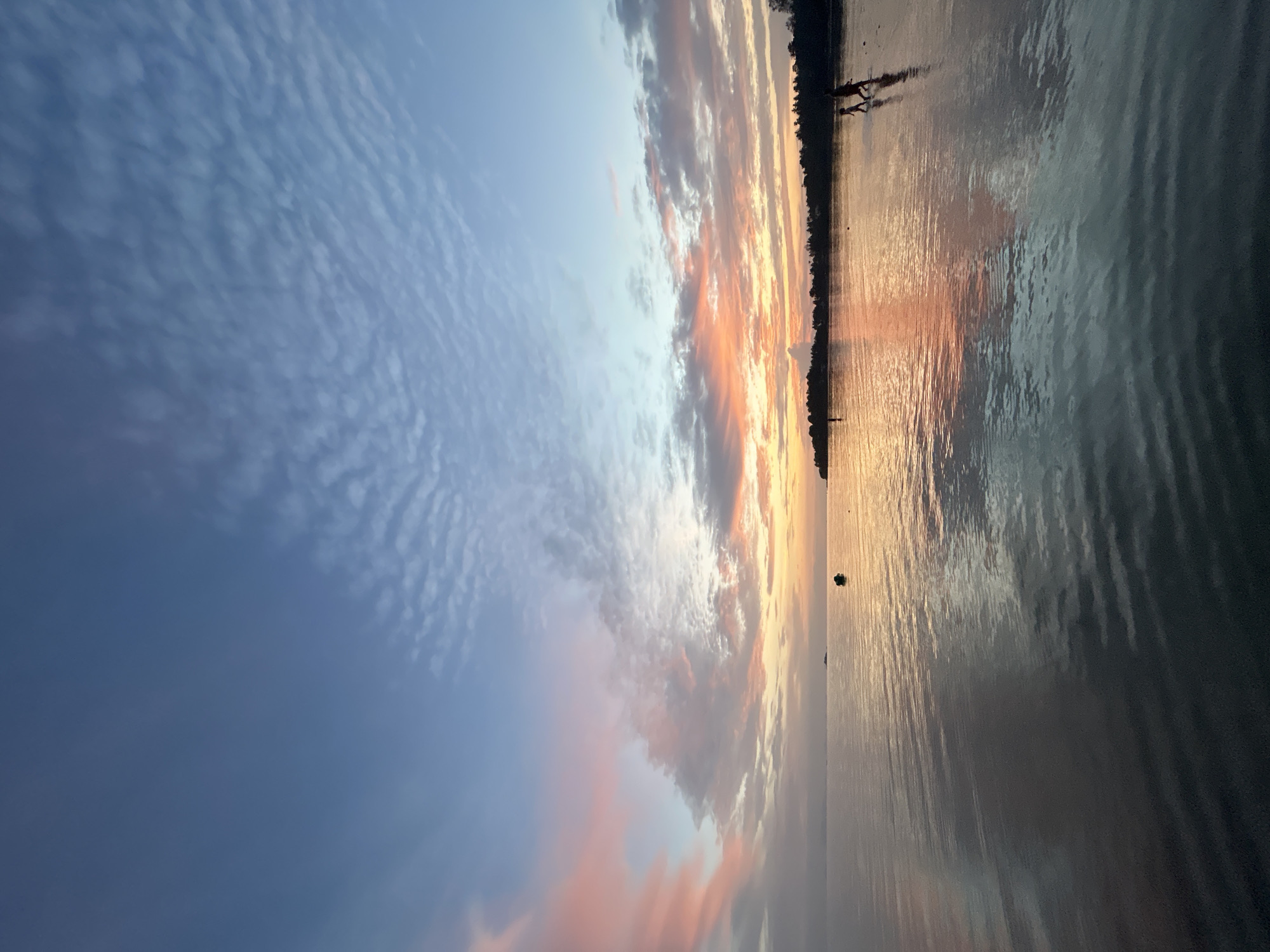 This image depicts a serene sunset scene at a beach in Ko Pha-ngan District, Thailand. The sky is a gradient of soft blues and pinks, with wispy clouds catching the last rays of sunlight. The calm water of the sea reflects the sky, creating a mirror-like surface with ripples. Along the horizon, a silhouette of lush vegetation and trees marks the coastline. In the foreground, two figures, appearing to be a child and an adult, are walking in the shallow water, their dark silhouettes stark against the illuminated water. A small boat is visible in the distance, anchored in the sea. The overall atmosphere is peaceful and tranquil, capturing the beauty of a tropical sunset.