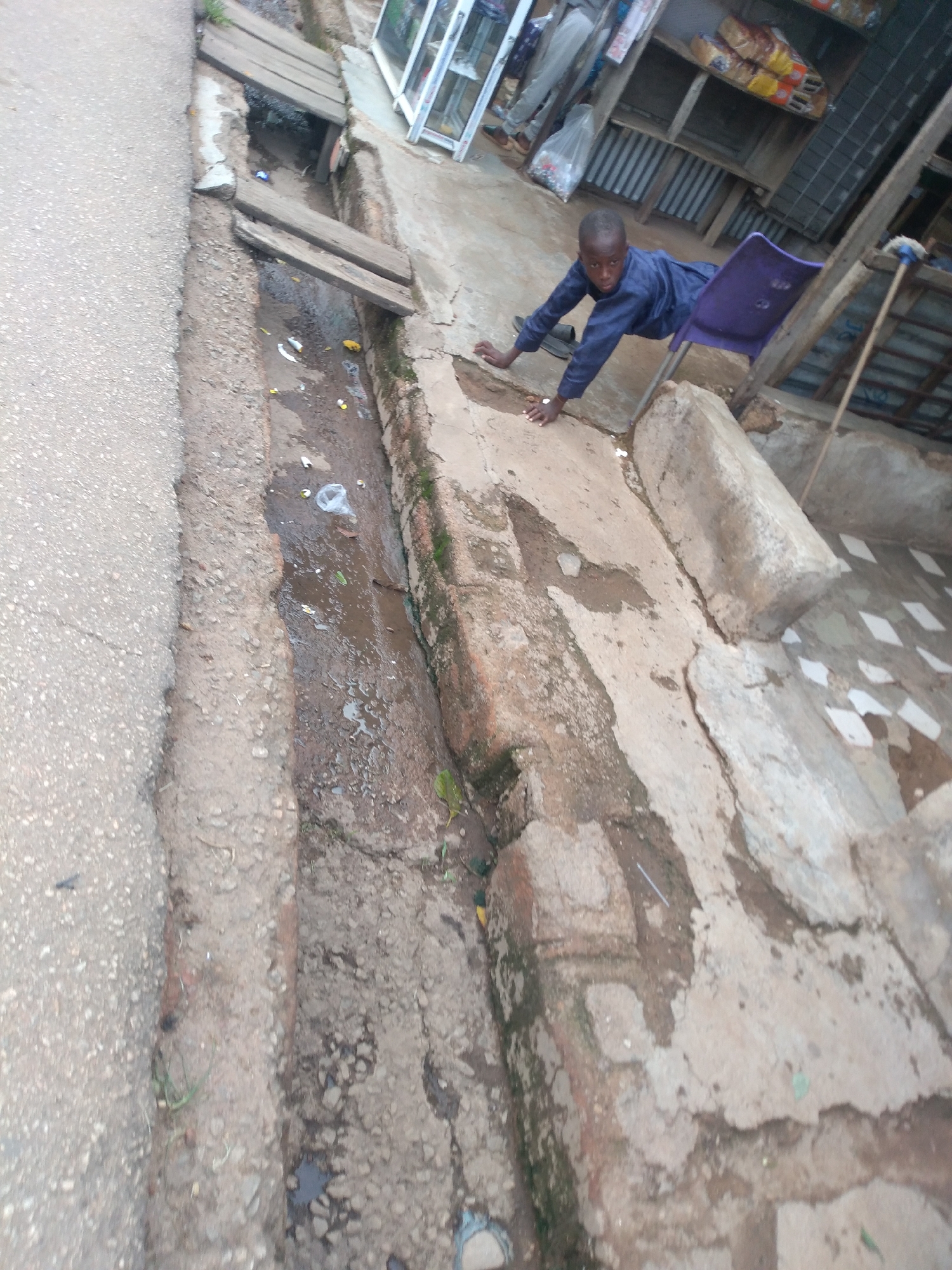 The image depicts a young boy, appearing to be of Nigerian descent, on his hands and knees on a cracked concrete surface next to a drainage ditch. He is wearing a dark blue long-sleeved shirt and dark trousers. A purple plastic chair is positioned behind him, and his left hand rests on it. To the left of the boy, a shallow, partially filled drainage ditch contains some debris and murky water. The ditch is bordered by concrete and dirt. In the background, a makeshift stall with wooden shelves is visible, stocked with various items, including what appears to be packaged bread. The overall environment suggests an outdoor, possibly urban, setting in a developing area. The presence of a drainage ditch and the visible stall could indicate a market or street-side commerce area.