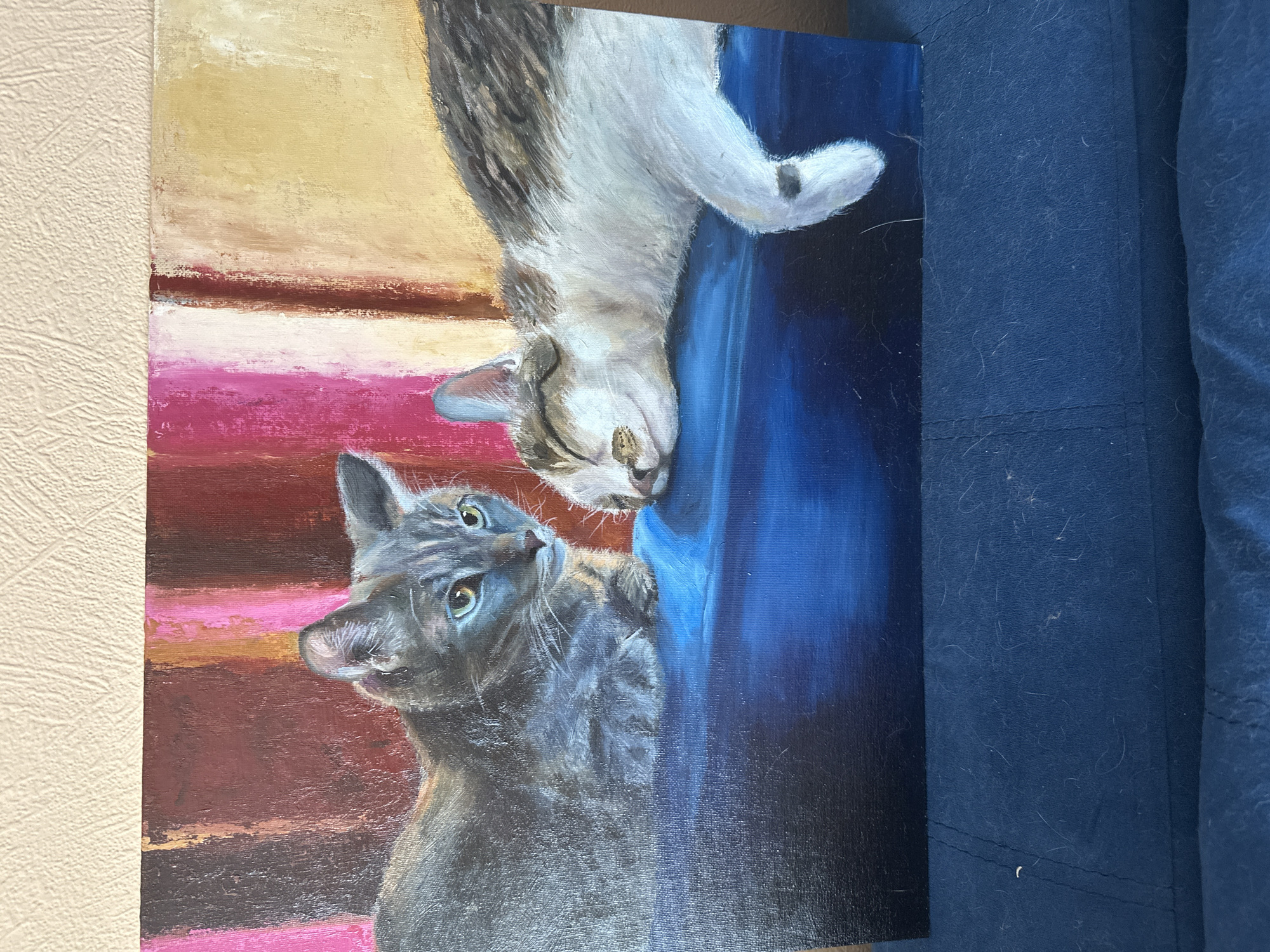 The image depicts a painting of two cats. One cat, a gray tabby, is looking up towards a second cat, a white and gray tabby, that is upside down. The background consists of a light yellow area at the top, a section with horizontal stripes of pink, white, and reddish-brown, and a deep blue area. The painting is placed next to a textured beige wall on the left and a dark blue fabric on the right. There is no indication that this image is located in Krasnoyarsk, Russia.