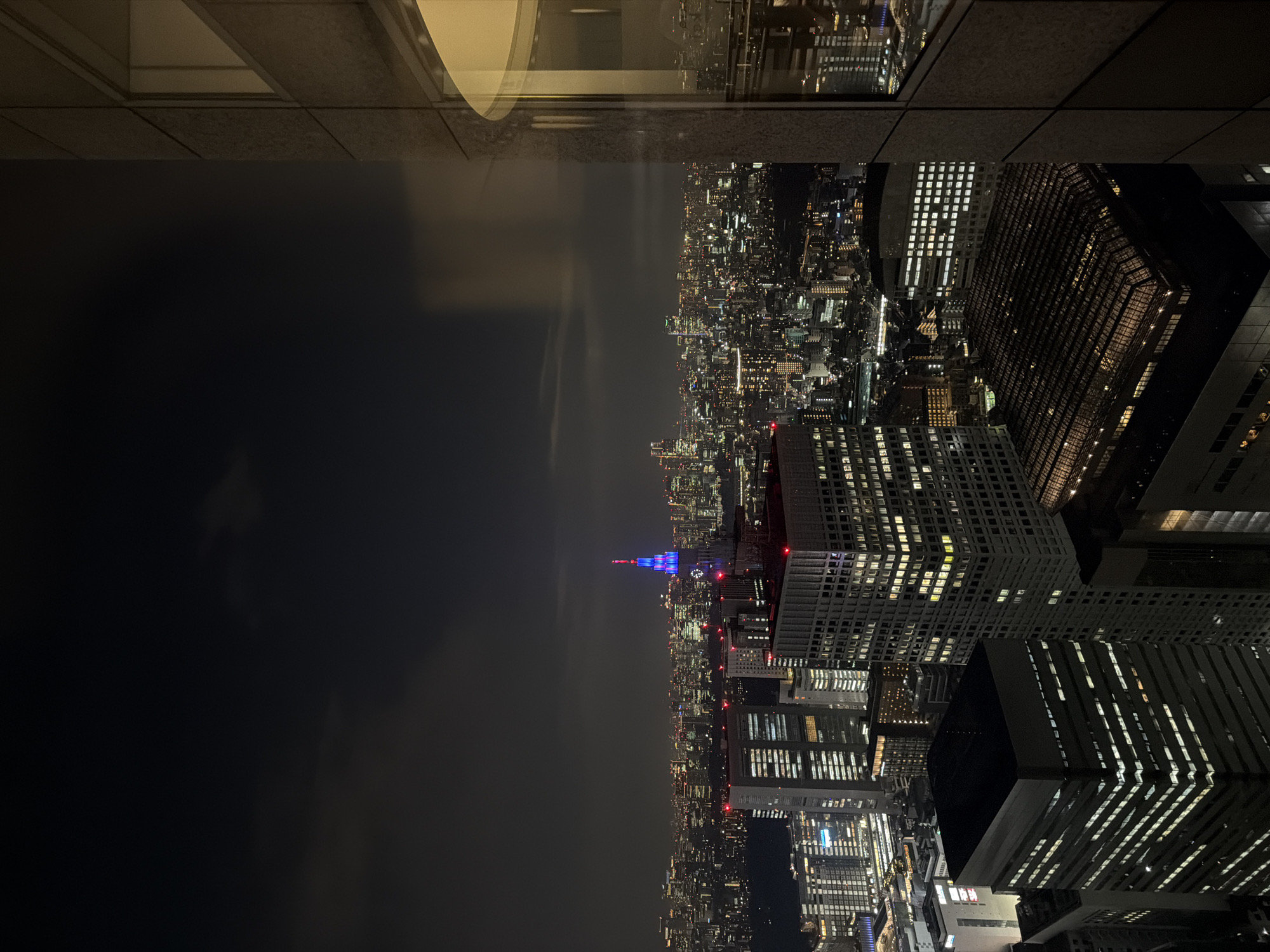 The image is a nighttime aerial view of the Shinjuku cityscape in Tokyo, Japan, taken from a high vantage point, likely a skyscraper. The scene is dominated by a dense concentration of illuminated skyscrapers and buildings, creating a sprawling urban panorama under a dark, cloudy sky. A prominent, tall building with a distinctive spire is illuminated with blue and red lights, standing out against the surrounding urban glow. The foreground is partially occupied by the corner of a modern building's exterior, featuring a large, curved glass window that reflects the distant city lights. The overall atmosphere is one of a vibrant, bustling metropolis at night, characterized by the extensive network of lights from countless windows and streetlights.