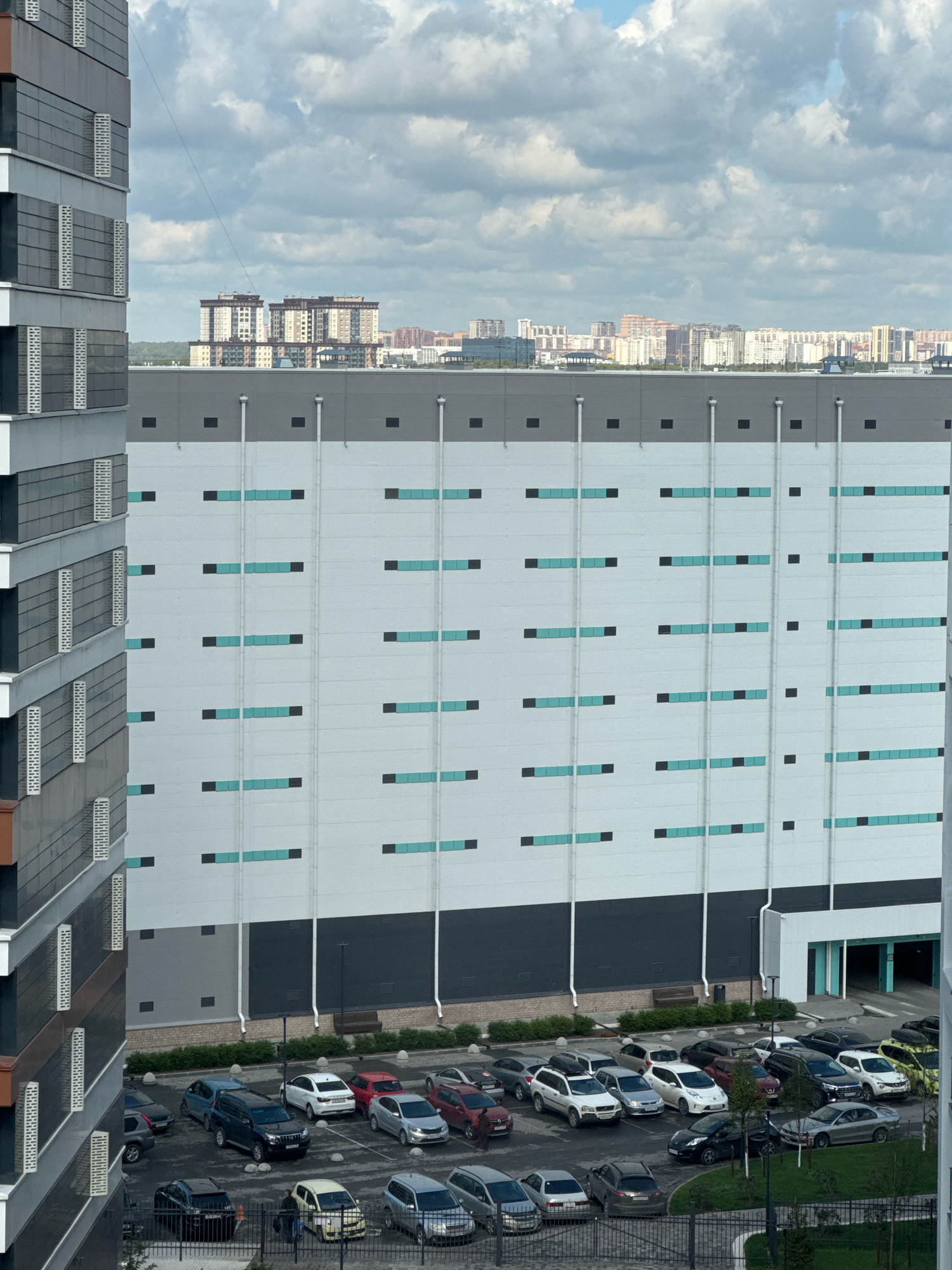 This is an aerial, eye-level photograph taken from a high vantage point, likely a residential building, looking towards a large, modern industrial or commercial building. The building on the right is vast and predominantly light gray with horizontal bands of turquoise and dark gray rectangles, likely decorative or functional elements. Vertical white pipes run down the facade. To the left of this large building is a tall, multi-story residential building with a facade of alternating dark gray and light gray rectangular panels and rows of white, grated vents or windows. The sky above is filled with heavy, gray clouds.
In the foreground and mid-ground, a paved parking lot is visible with numerous cars parked. The cars are of various makes and models, including SUVs, sedans, and hatchbacks, in colors such as white, silver, black, red, and blue. Some people are visible in the parking lot, appearing to be walking or standing near vehicles. A dark metal fence runs along the bottom edge of the frame, separating the parking lot from a landscaped area with bushes and trees. In the distance, a cityscape is visible with a cluster of multi-story residential buildings under the cloudy sky.
Based on the architectural style and the context of similar structures, this scene is likely located in a modern urban development area in Novosibirsk, Russia. The specific location appears to be near a logistical or industrial park situated adjacent to a residential complex. The time of day seems to be late afternoon or early evening, given the diffused natural light and the cloud cover.
