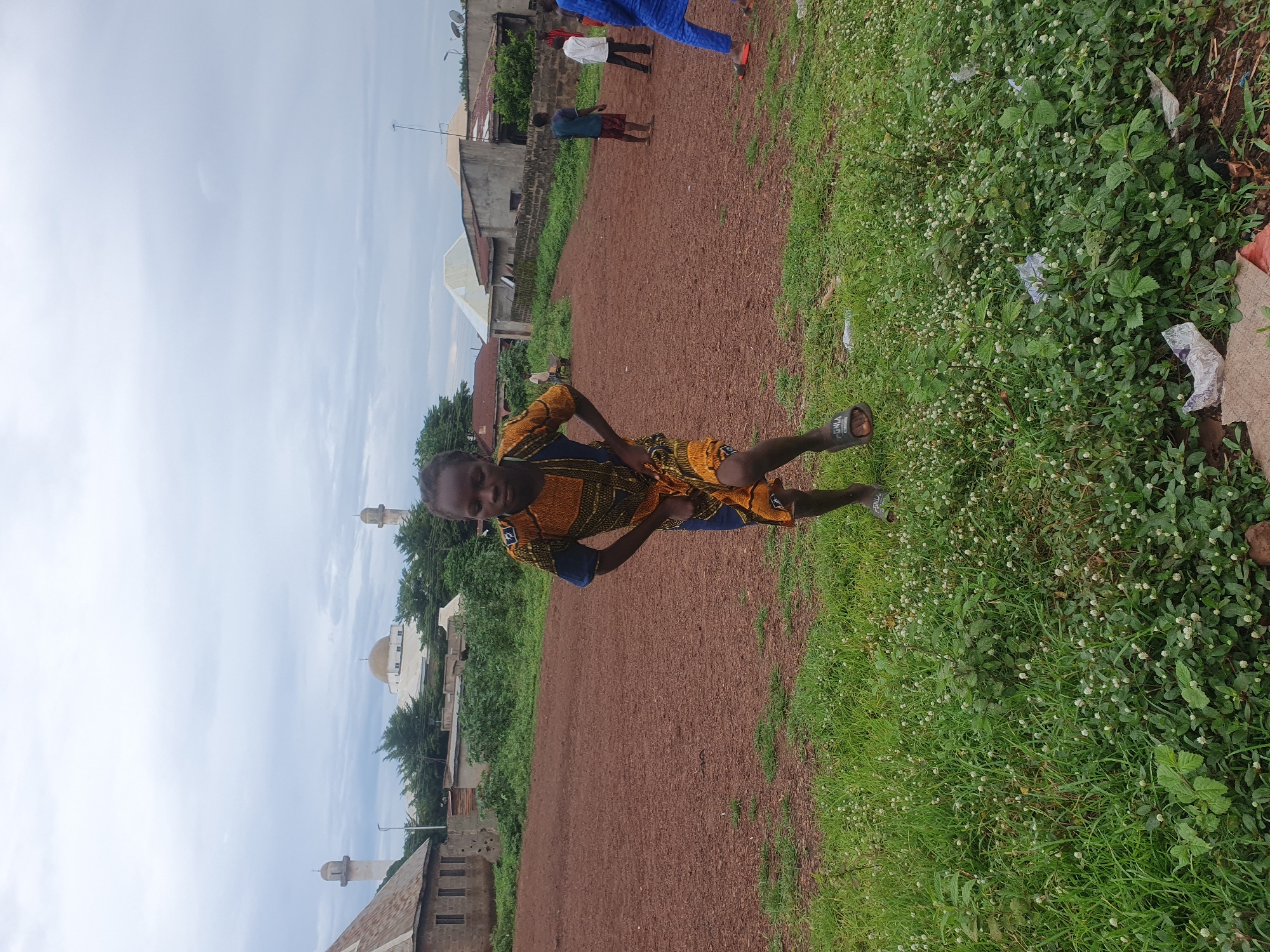 This image captures an outdoor scene in Idah, Nigeria, under an overcast sky. The foreground features a young individual with dark skin, wearing a patterned top and shorts in yellow, blue, and brown hues, along with dark sandals. This individual is positioned centrally, facing the viewer with knees bent and hands placed on the hips or upper thighs, suggestive of a pose or movement.
Behind the foreground subject, a vast, reddish-brown dirt field dominates the midground, interspersed with patches of green vegetation. Several other individuals, appearing to be children or young adults, are visible in the distance on the right side of the field. One wears a dark shirt and reddish shorts, another a white shirt and dark pants, and a third a vibrant blue traditional garment with orange flip-flops. They appear to be standing or walking.
The background reveals an array of structures. To the far left, a building with a reddish-brown roof and exposed block walls is visible. Towards the center of the background, a large white mosque with a prominent golden dome and two tall, slender minarets topped with smaller domes stands out against the sky. Two additional minarets, similar in design but partially obscured, are visible further left and right. Other residential or commercial buildings with various roof styles and satellite dishes are scattered across the background.
The immediate foreground consists of lush green grass dotted with numerous small white wildflowers. Scattered pieces of white plastic debris are visible within this grassy area, particularly on the lower right. The sky is uniformly light grey, indicating heavy cloud cover.
