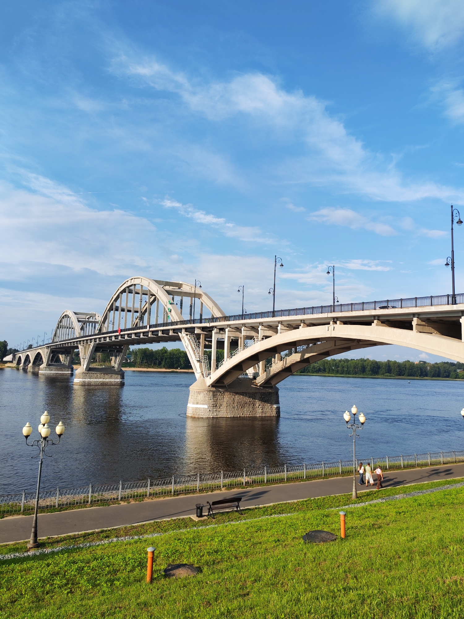 The image shows a wide, concrete arched bridge spanning a calm river under a bright blue sky with scattered white clouds. The bridge has multiple arches and is supported by stone pillars. Along the riverbank in the foreground, there is a paved walkway with lampposts and a metal fence. Green grass covers the embankment. A few people are walking on the path, and there is a bench and a trash can near the walkway. Trees line the opposite bank of the river. The scene appears to be a picturesque view of a city park or waterfront area in Bol'shie Mkhi, Russia.