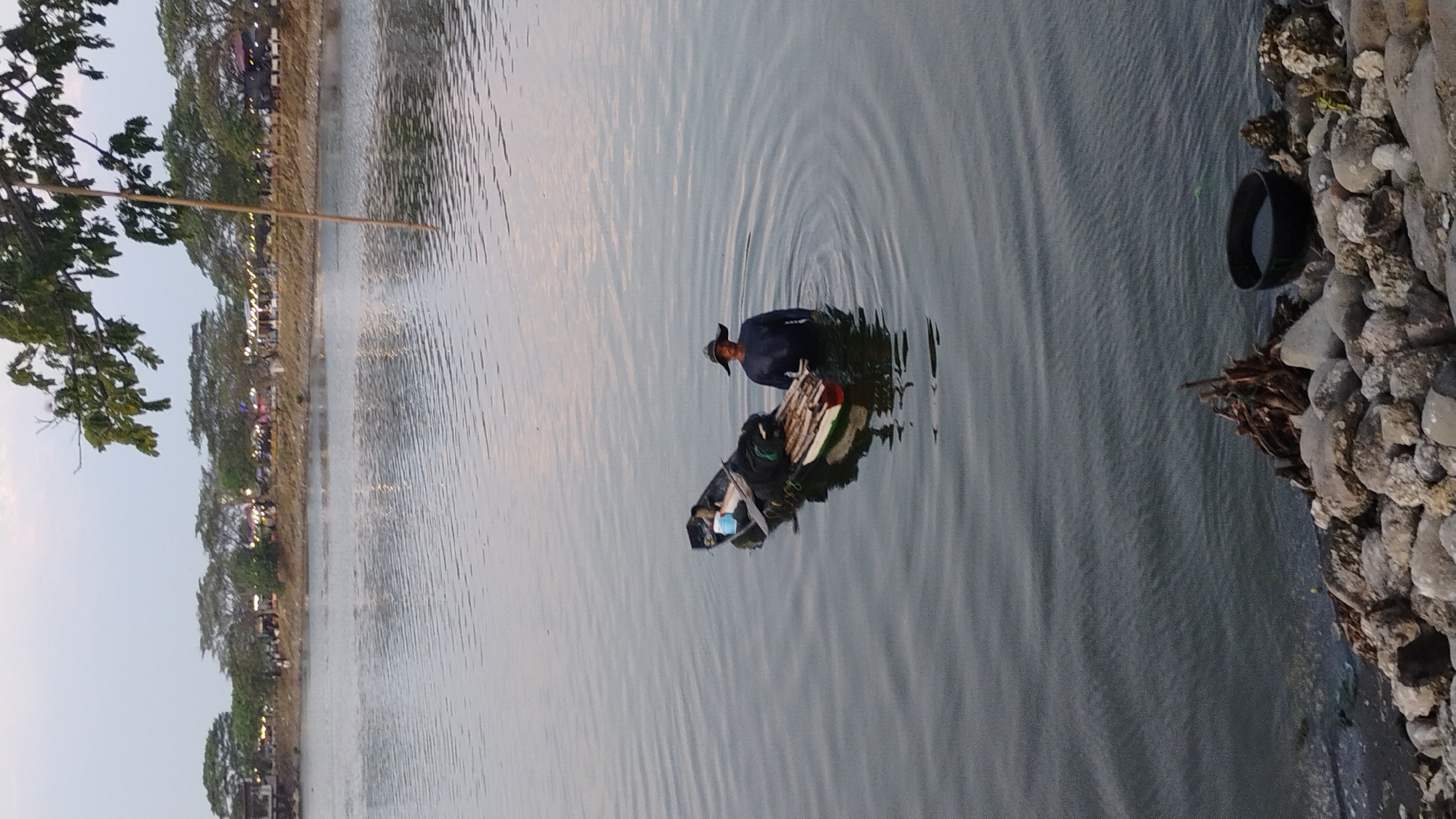 The image depicts a man standing in shallow water next to a small boat loaded with various items. The boat appears to be a traditional Indonesian fishing vessel, with a long, narrow hull. The man is wearing a dark long-sleeved shirt and a wide-brimmed hat. He is positioned in the water between the boat and the shore. The boat contains bundles of wood or bamboo, nets, and other supplies. The water is calm with ripples spreading outwards from the boat.
In the background, the shore is lined with trees and some structures, possibly shops or restaurants, with lights visible indicating it might be evening. The sky is overcast with a soft, diffused light. The foreground shows a rocky shoreline with some driftwood.
This scene is likely located at a recreational area or a fishing spot along the coast or a lake in Lhokseumawe, Indonesia. The presence of the boat, fishing gear, and the surrounding environment are consistent with a coastal or lakeside community in Indonesia.