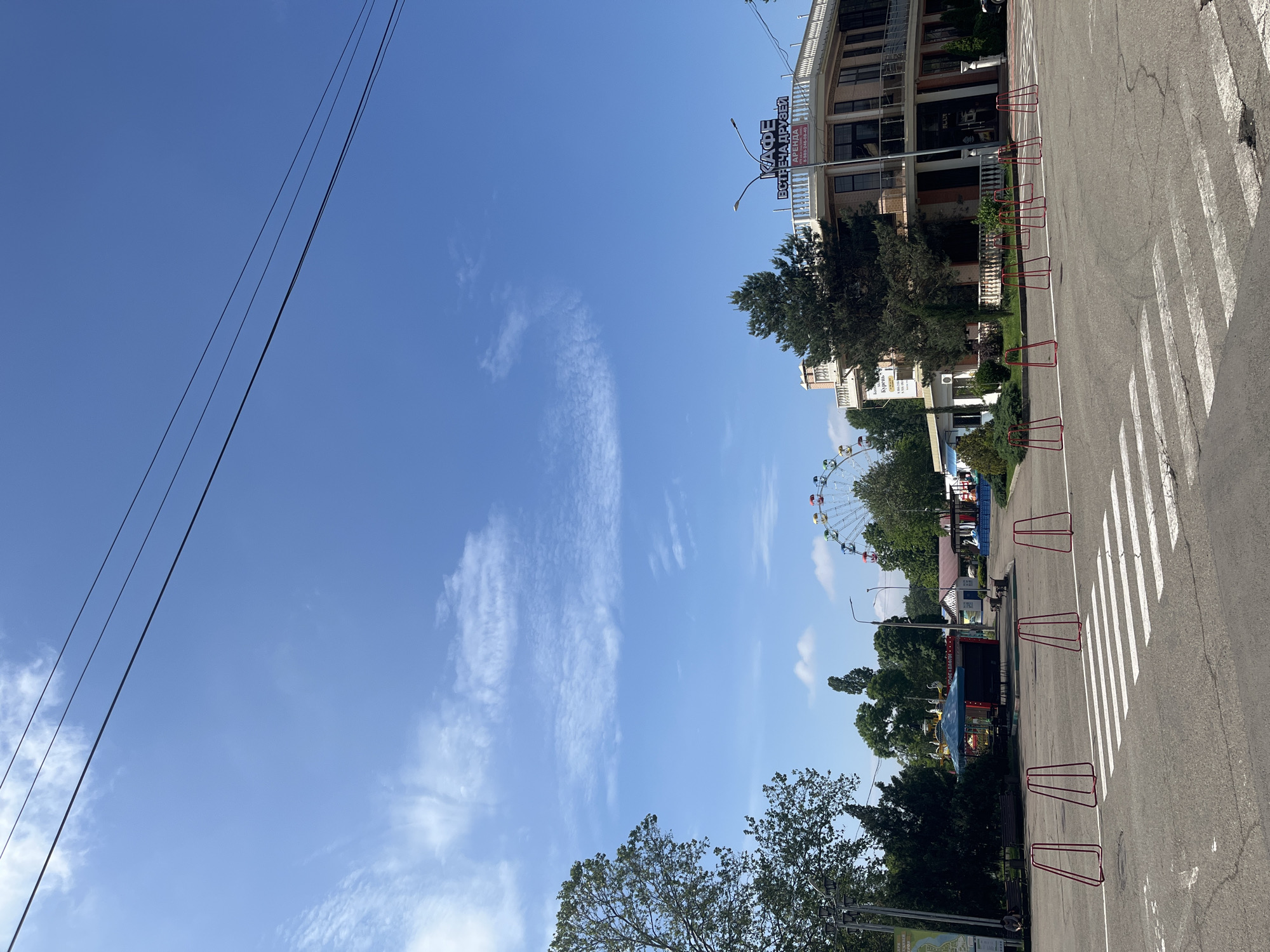 The image depicts an outdoor scene under a clear blue sky with scattered white clouds, suggesting a sunny day, likely mid-morning to late afternoon. The perspective is looking down a street or paved path on the right, which features a zebra crossing and several red metal barriers placed along its edge.
On the right side of the frame, a multi-story building with balconies and windows is prominent. A large commercial sign is mounted on its upper facade, displaying text in Cyrillic. Further down the path, surrounded by trees, a colorful Ferris wheel is visible in the distance, indicating the presence of an amusement park or fairground. Lush green trees line the street and are scattered throughout the background, providing shade.
Across the upper left portion of the sky, two dark power lines stretch diagonally. No people are visible, and no specific activity is taking place, giving the scene a calm and still atmosphere.
Visible text on the commercial sign reads "КАФЕ ВСТРЕЧА ДРУЗЕЙ", which translates to "CAFE MEETING OF FRIENDS". Below this, smaller text reads "АРЕНДА" (RENT) and a phone number "8-989-240-59-40". Another sign further down the street is partially visible with the word "hyper". Given the location context, this scene is in Krasnodar, Russia.