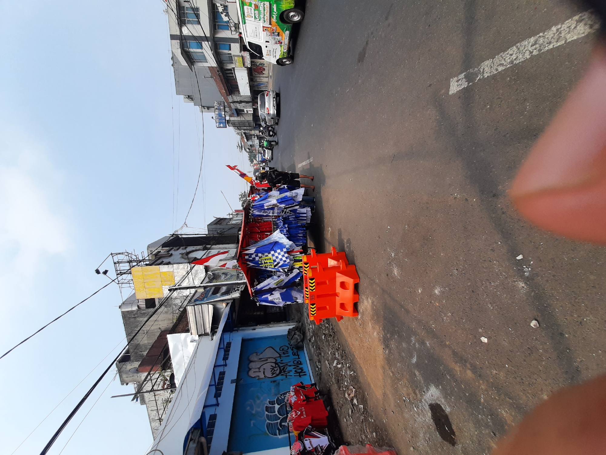 The image shows a street scene in Bandung City, Indonesia, with a focus on a vendor stall selling merchandise on the side of the road. The stall, a portable cart decorated with flags, displays numerous blue and white checkered scarves and other fabric items, likely fan merchandise. In front of the stall are several bright orange plastic barriers, commonly used for traffic control or to delineate pedestrian areas. To the left of the stall, red t-shirts with black lettering are displayed on a rack, with a blue wall featuring graffiti behind them. The street itself is paved, with traffic visible in the background, including cars and motorcycles. A white van with colorful advertisements is parked on the right side of the road. The buildings lining the street are a mix of older and newer structures, some appearing to be shops or commercial establishments. The sky is clear and bright, indicating daylight. Two individuals are visible near the stall, one standing and one seated, appearing to be engaged with the merchandise or the stall itself. The overall environment suggests a bustling urban street with commercial activity.