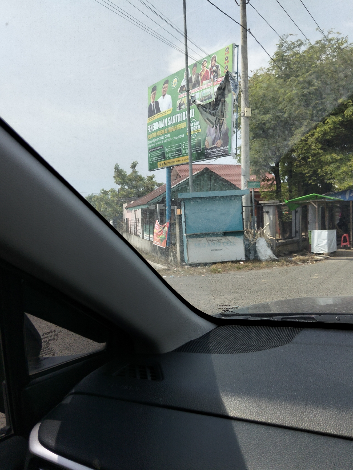 The image shows a street scene in Geulanggang Teungoh, Indonesia, viewed from inside a vehicle. In the foreground, the dashboard of the car is visible, along with a portion of the windshield reflecting the environment outside. A large green billboard is prominently displayed on the right side of the road, featuring text in Indonesian and images of people, advertising "PENERIMAAN SANTRI BARU" (New Student Admissions) for "PESANTREN MODERN AL ZAHRAH BIRU EUEN" (Al Zahrah Biru Euen Modern Islamic Boarding School). The billboard appears weathered and partially torn. Behind the billboard, trees and buildings can be seen. To the left of the billboard, a small building with a blue facade stands. There are also some stalls or kiosks along the road. A utility pole with wires runs across the top of the image. The environment appears to be outdoors with daylight.