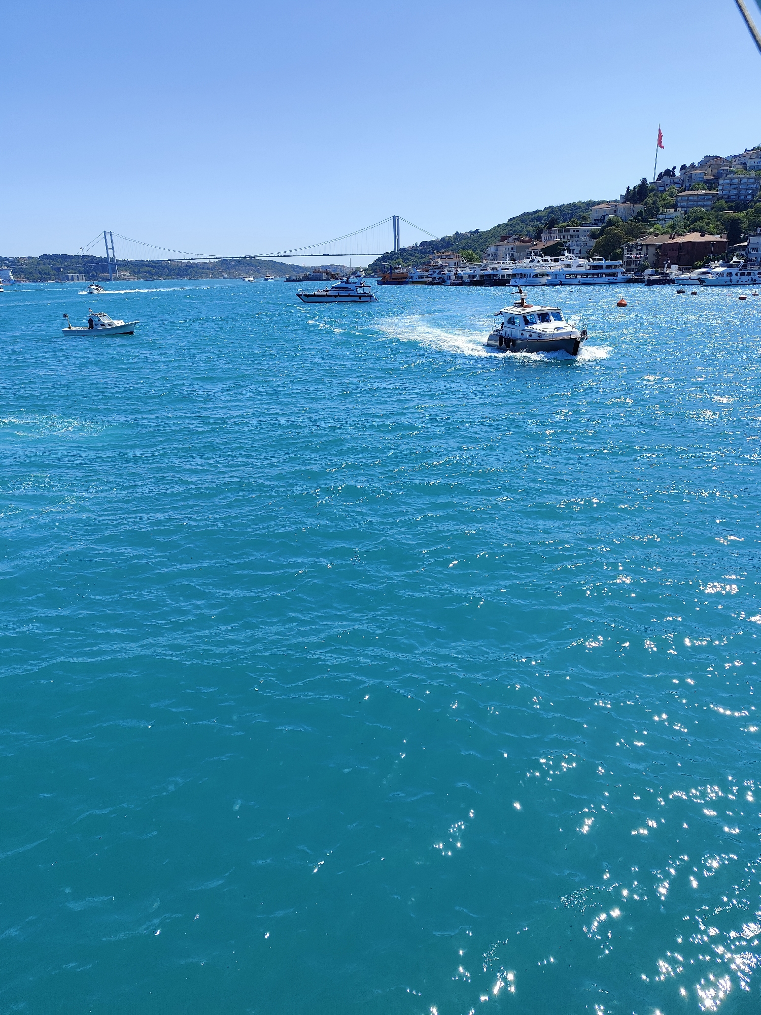 The image depicts a maritime scene on the Bosphorus in Beşiktaş, Turkey on a sunny day. The camera is positioned at an elevated perspective overlooking the water. The water is a bright turquoise color with sun glints, and several boats are visible. In the foreground, a small motorboat is cutting through the water, creating a wake. To the left, a smaller, open boat with at least one person on board is visible. In the middle distance, a larger ferry moves across the frame. The background features the shoreline with buildings and vegetation. The Bosphorus Bridge (15 July Martyrs Bridge) spans the horizon. The scene appears to be a common view for those in the area of the Ortaköy neighborhood.